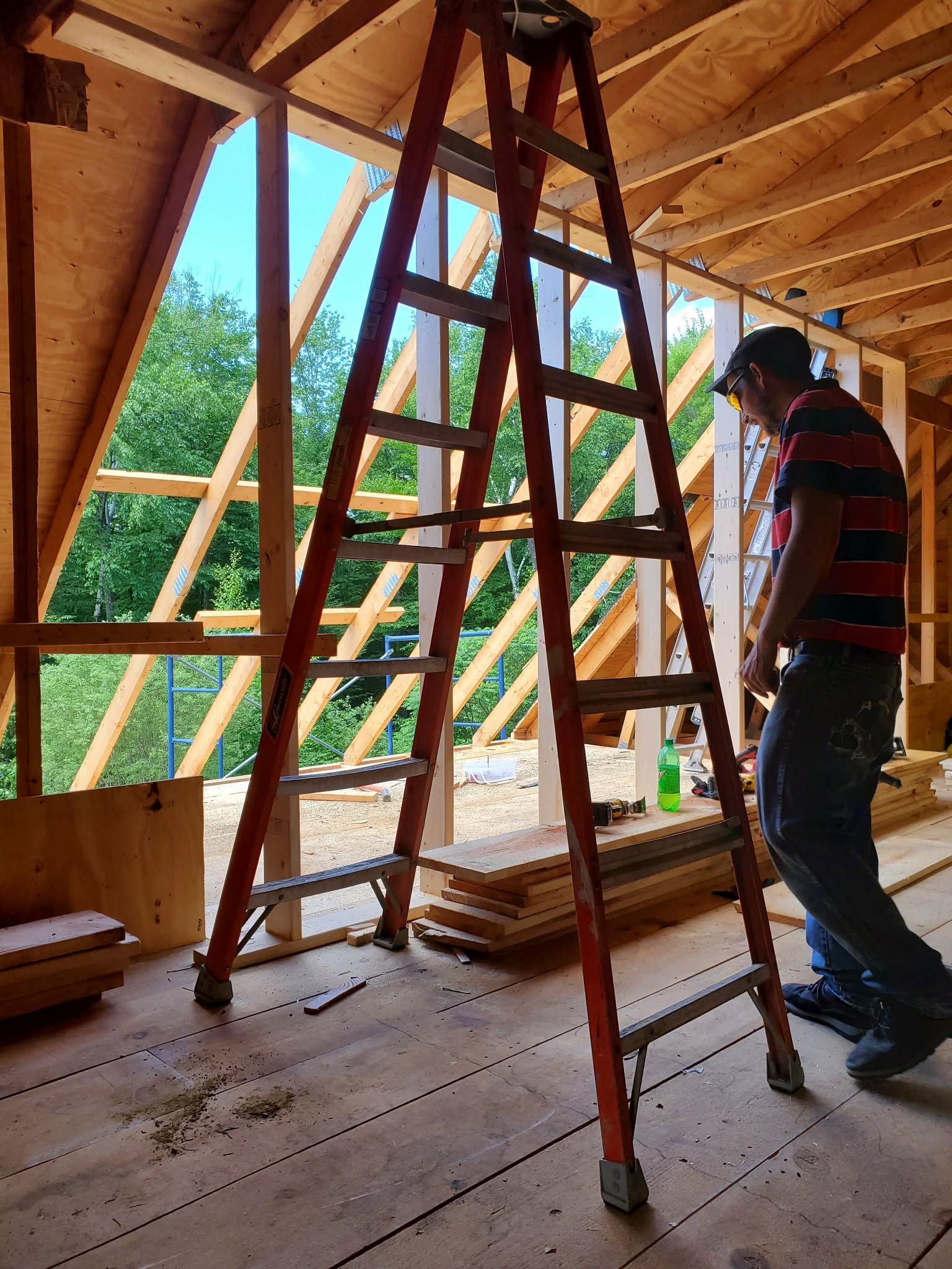 Person measuring inside a wooden house frame beside a ladder, with exposed roof beams and window openings