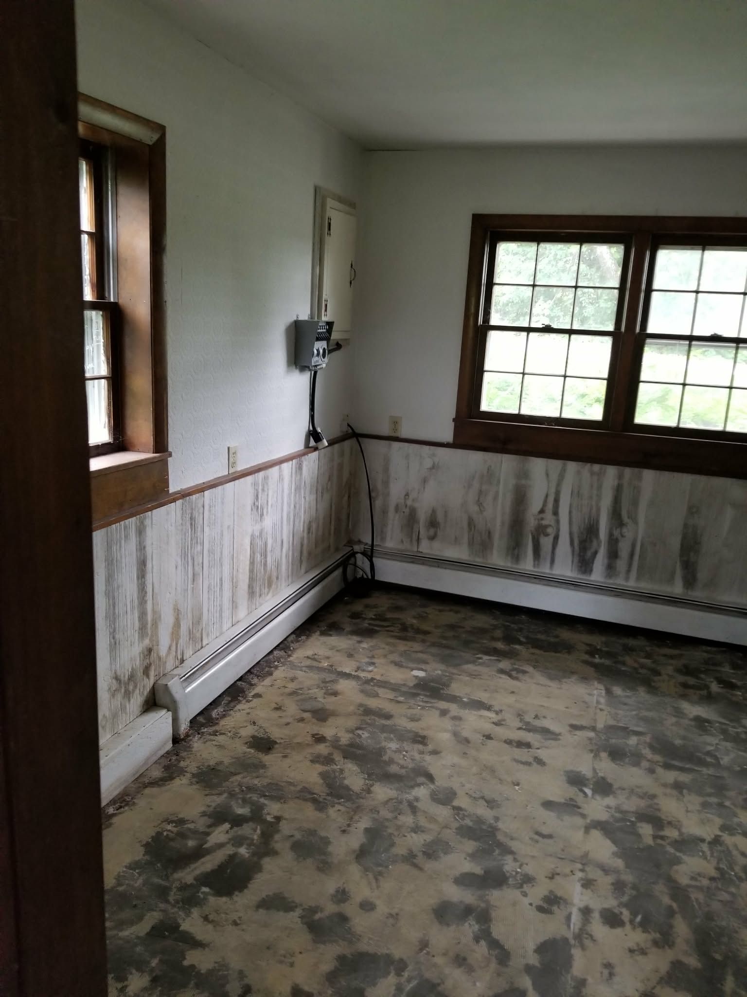 Empty, worn room with peeling walls, two windows, and a damaged floor.