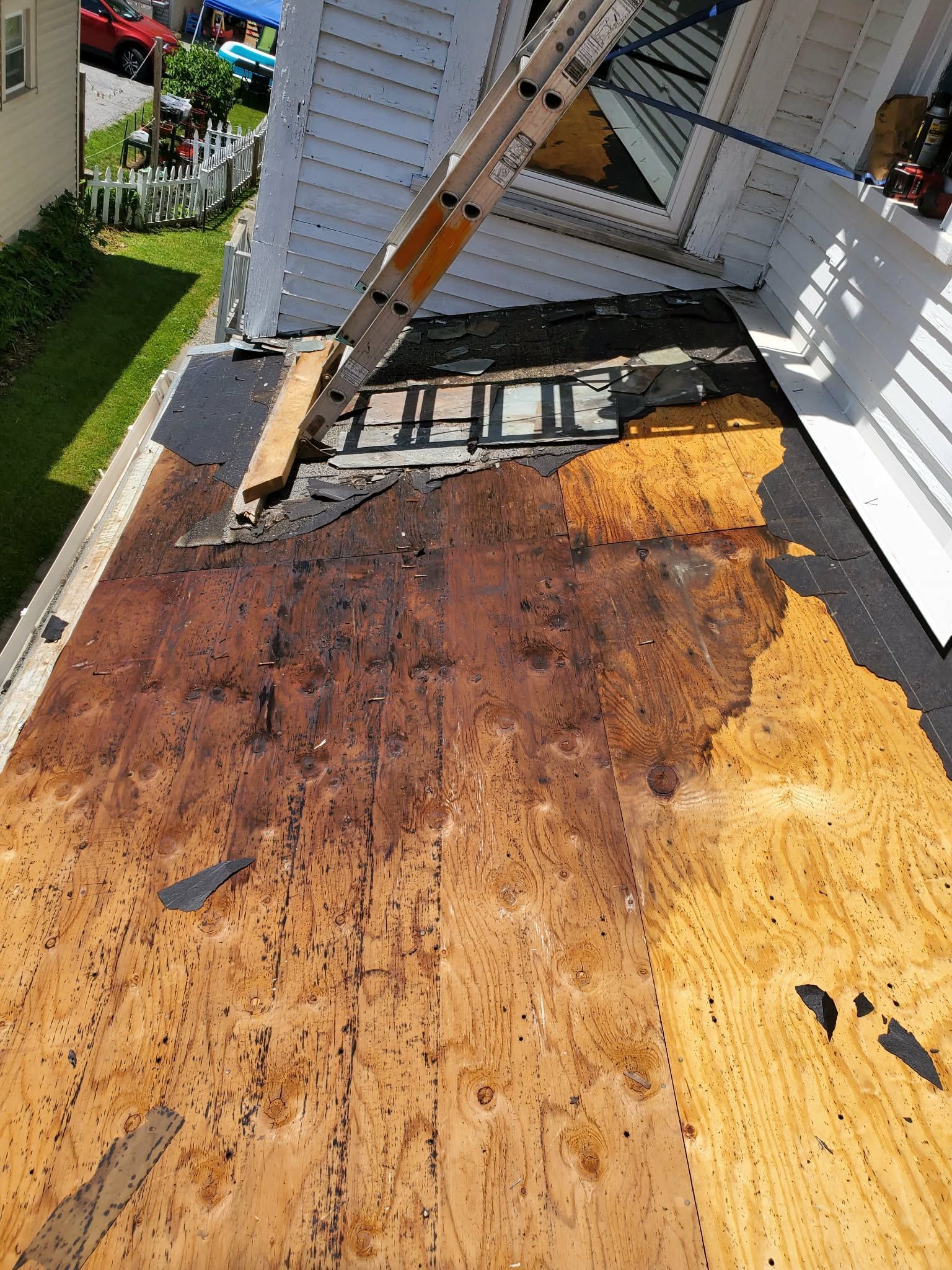 Rusted metal roof being removed, exposing plywood decking beside a white house and ladder.