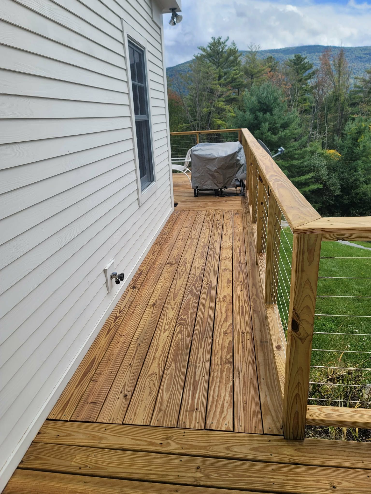 Wooden deck beside a house, with railing and a covered grill overlooking trees and mountains.