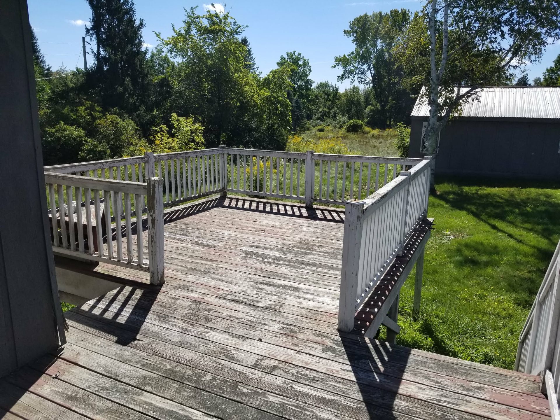 Weathered wooden deck with white railings overlooking a grassy yard and trees under a bright sky