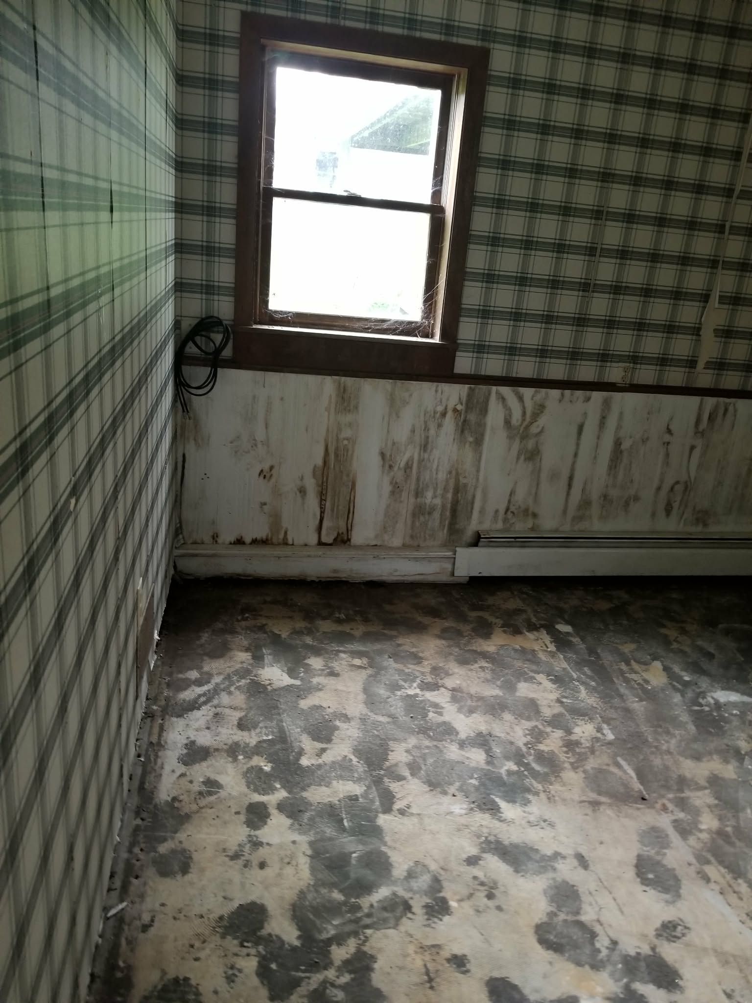Small empty room with peeling white walls, checkered tile wall, stained floor, and one window.
