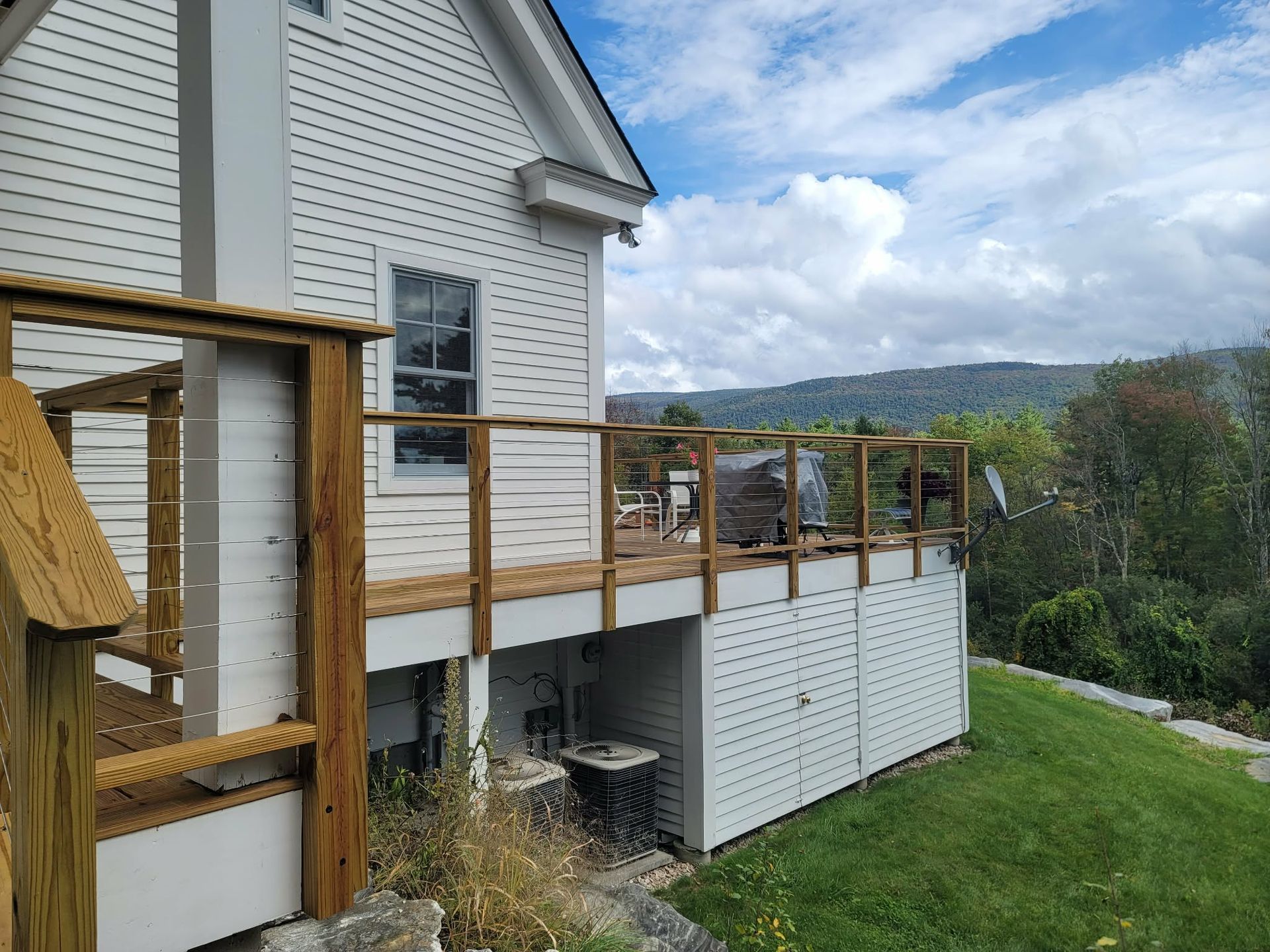 White house with wooden deck and railing overlooking a grassy yard and wooded hillside.