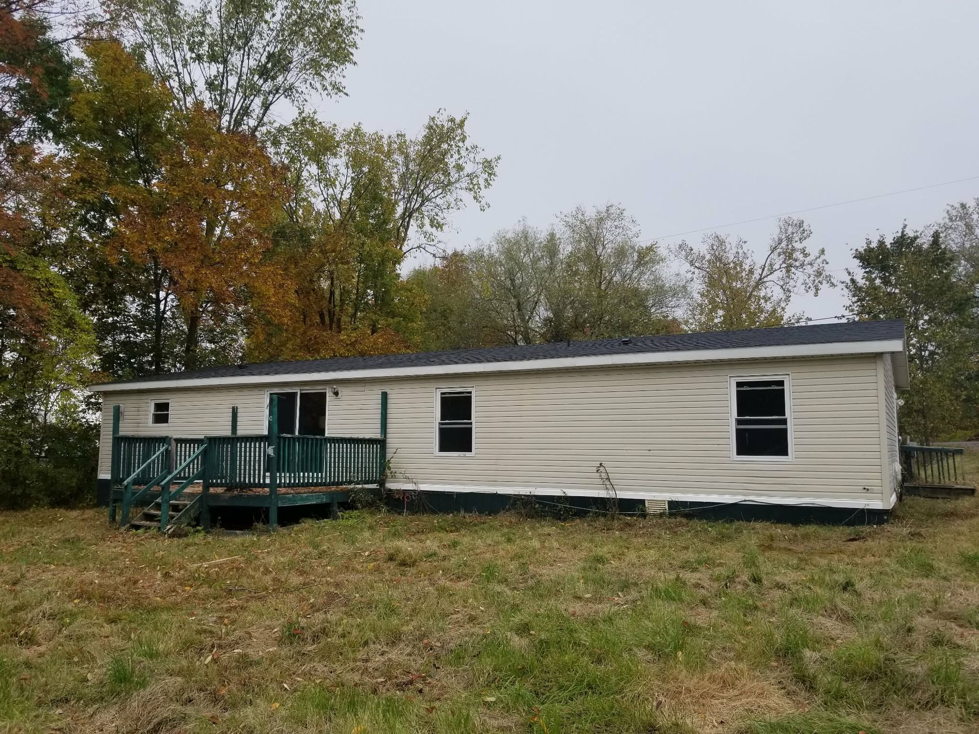 White single-wide trailer with green porch on a grassy lot beside autumn trees