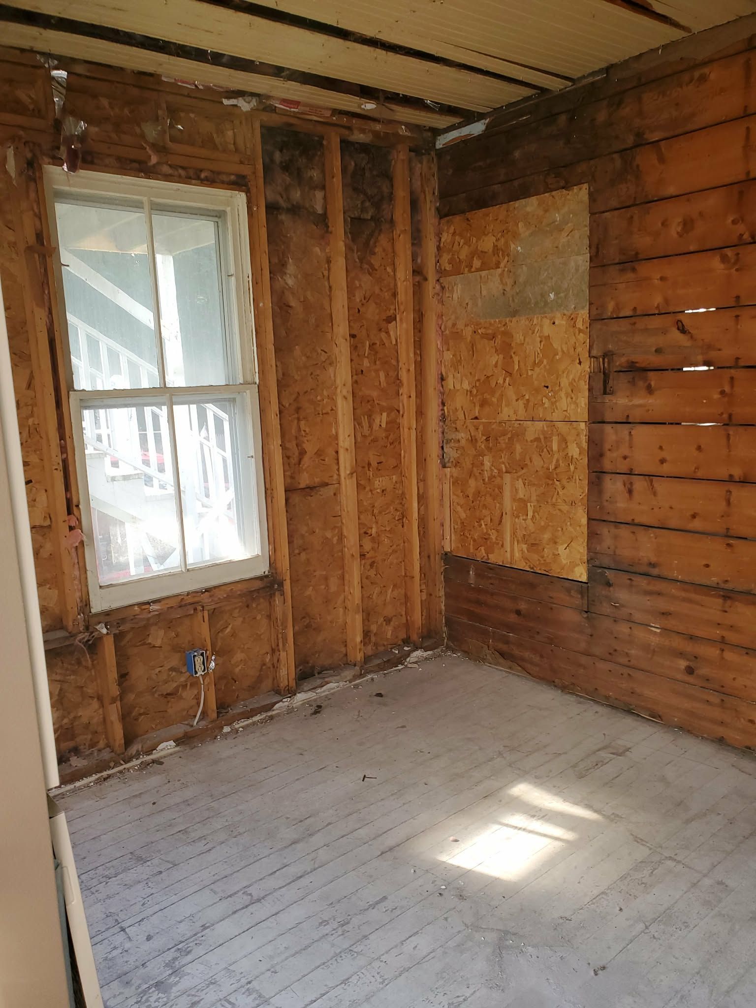 Bare, unfinished room with exposed wood framing, a window, plywood walls, and sunlight on the floor.