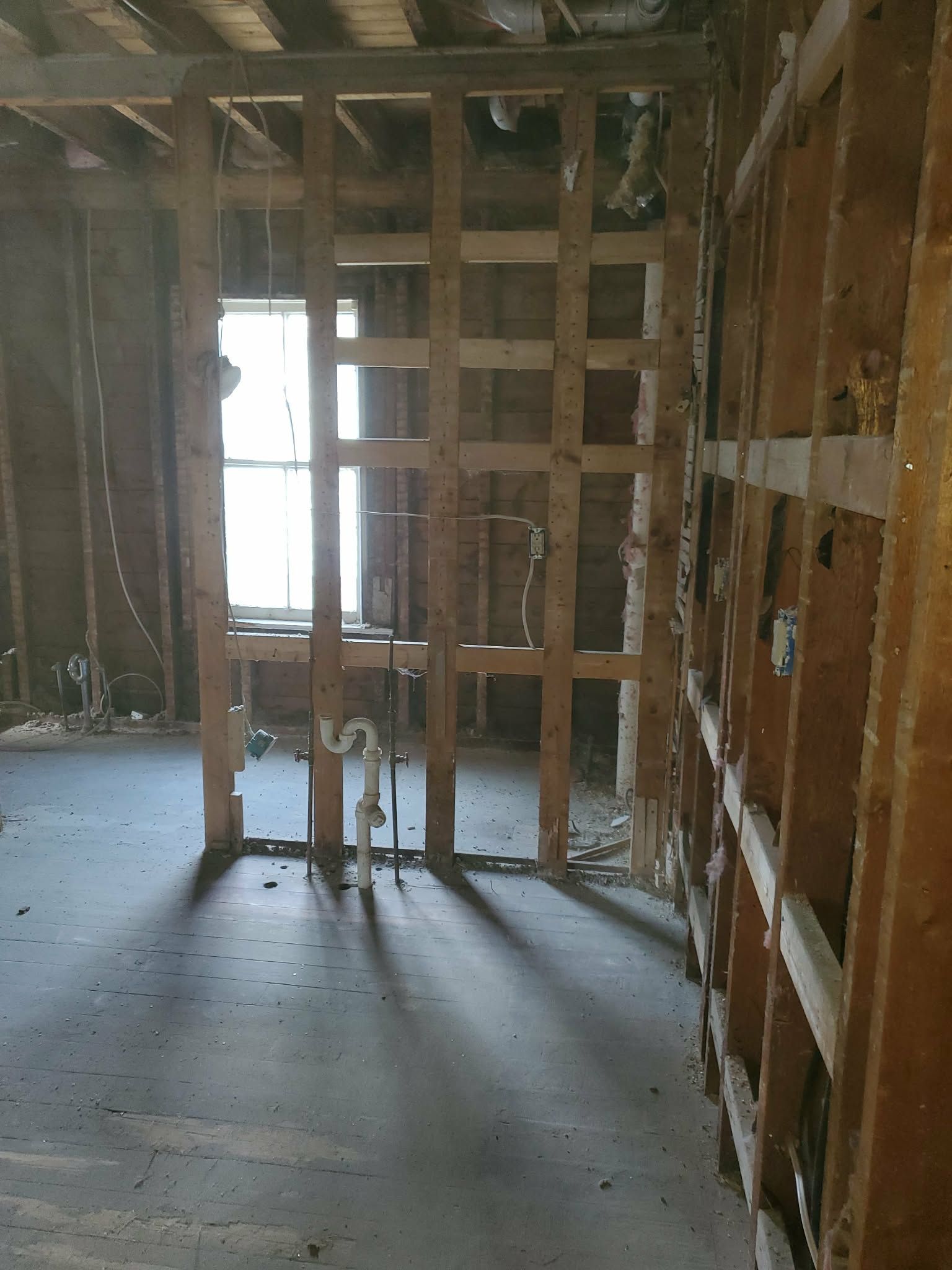 Interior under renovation with exposed wall framing, unfinished drywall, and light from a window at the back.