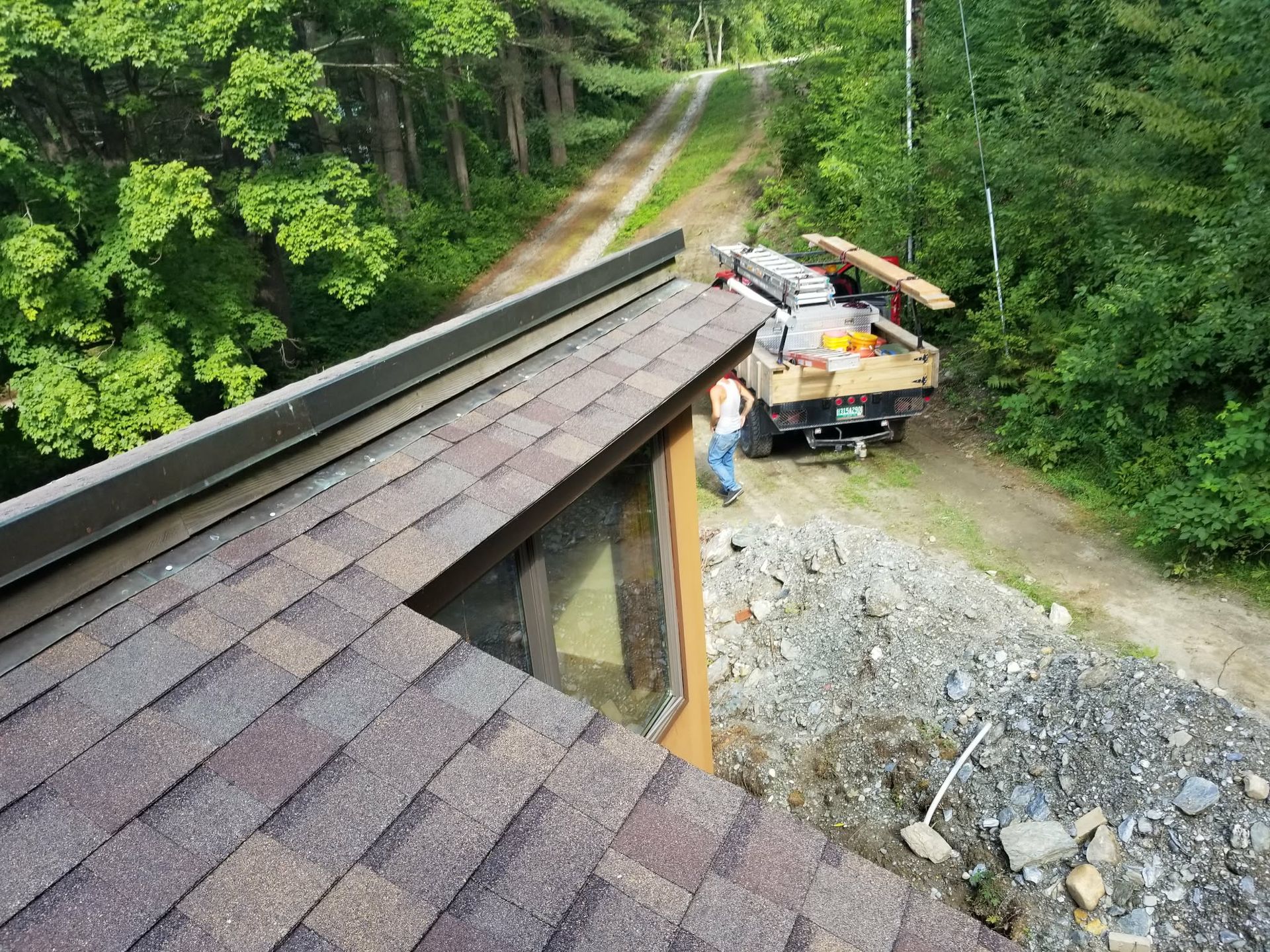 Roofing materials and a truck on a steep cabin roof beside a gravel road in a wooded area