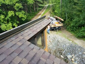 Roofing work on a cabin roof beside a gravel road in a wooded area