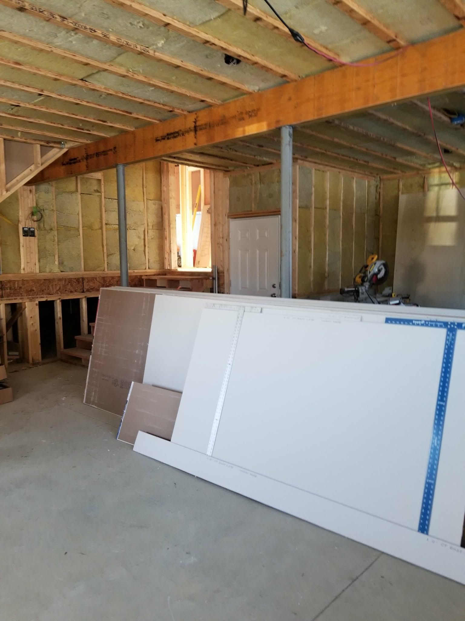 Unfinished room with exposed framing and insulation, leaning drywall sheets, and a concrete floor.