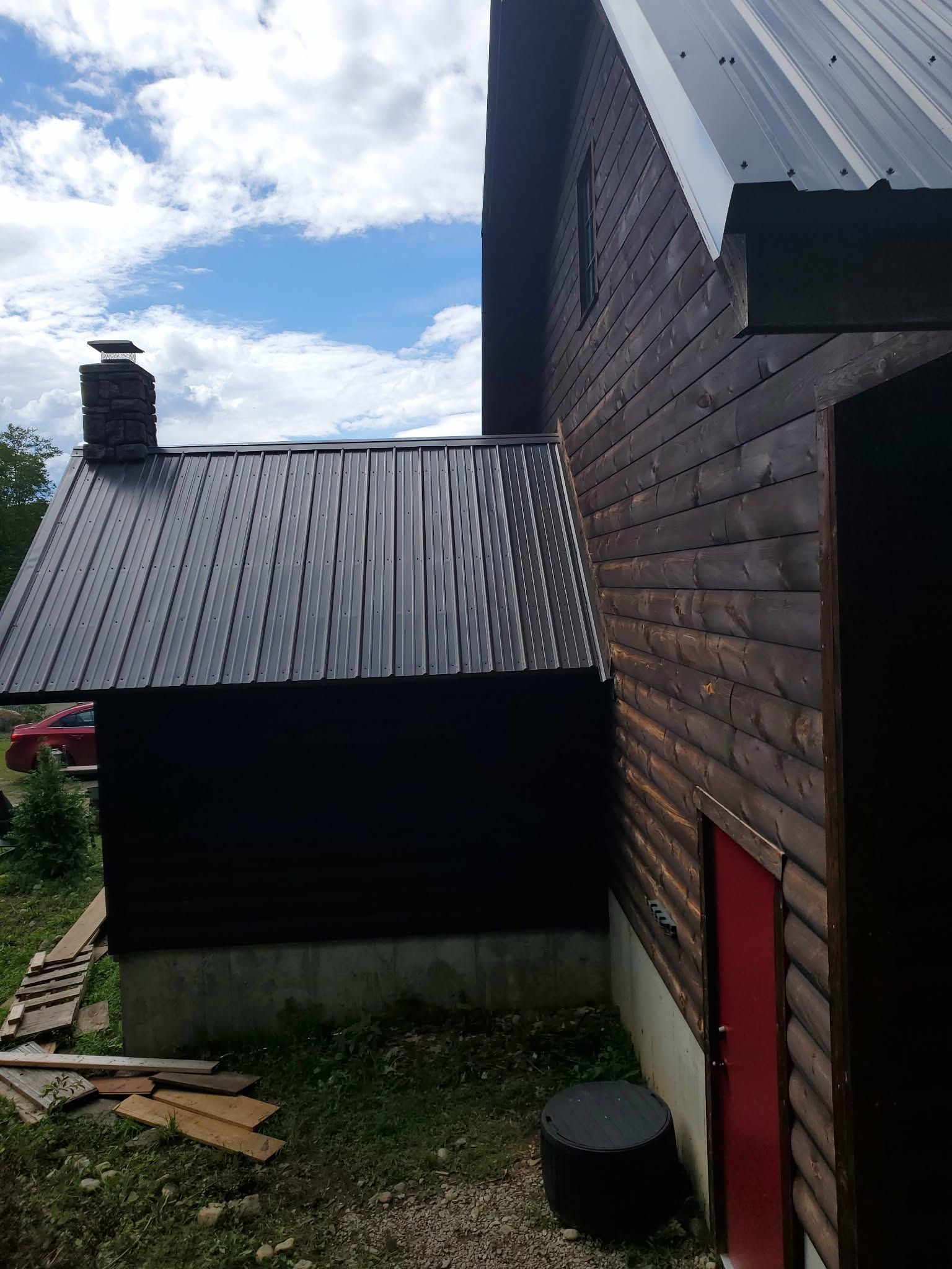 Wooden house exterior with corrugated metal roofs, a red door, and a grassy yard under a cloudy sky