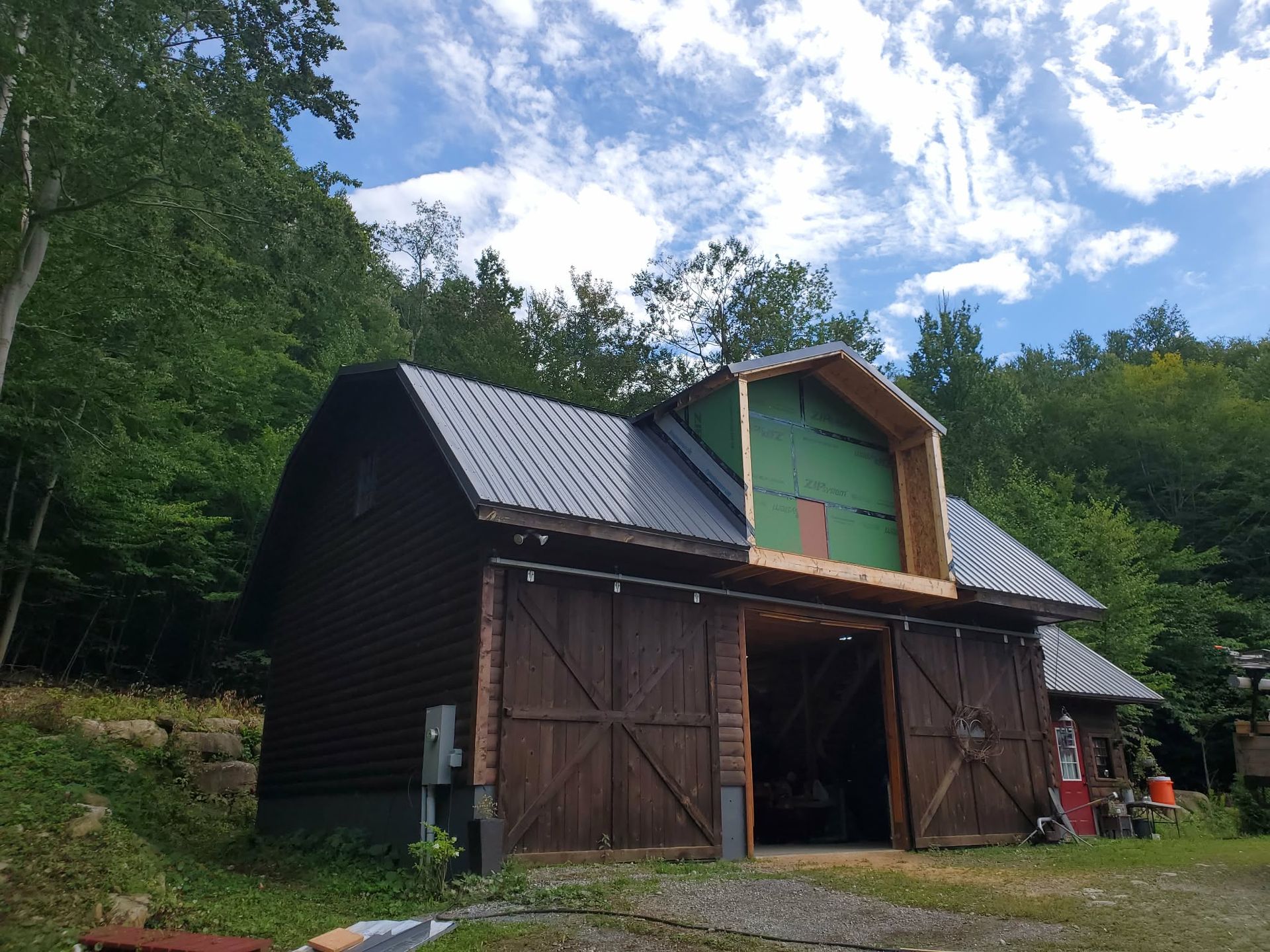 Barn with dark siding and large wooden doors in a wooded clearing under a cloudy sky