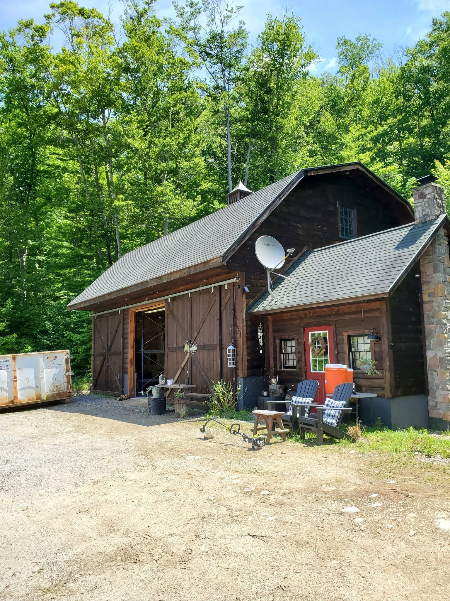Rustic brown cabin in a sunny wooded clearing with a gravel lot and outdoor chairs.