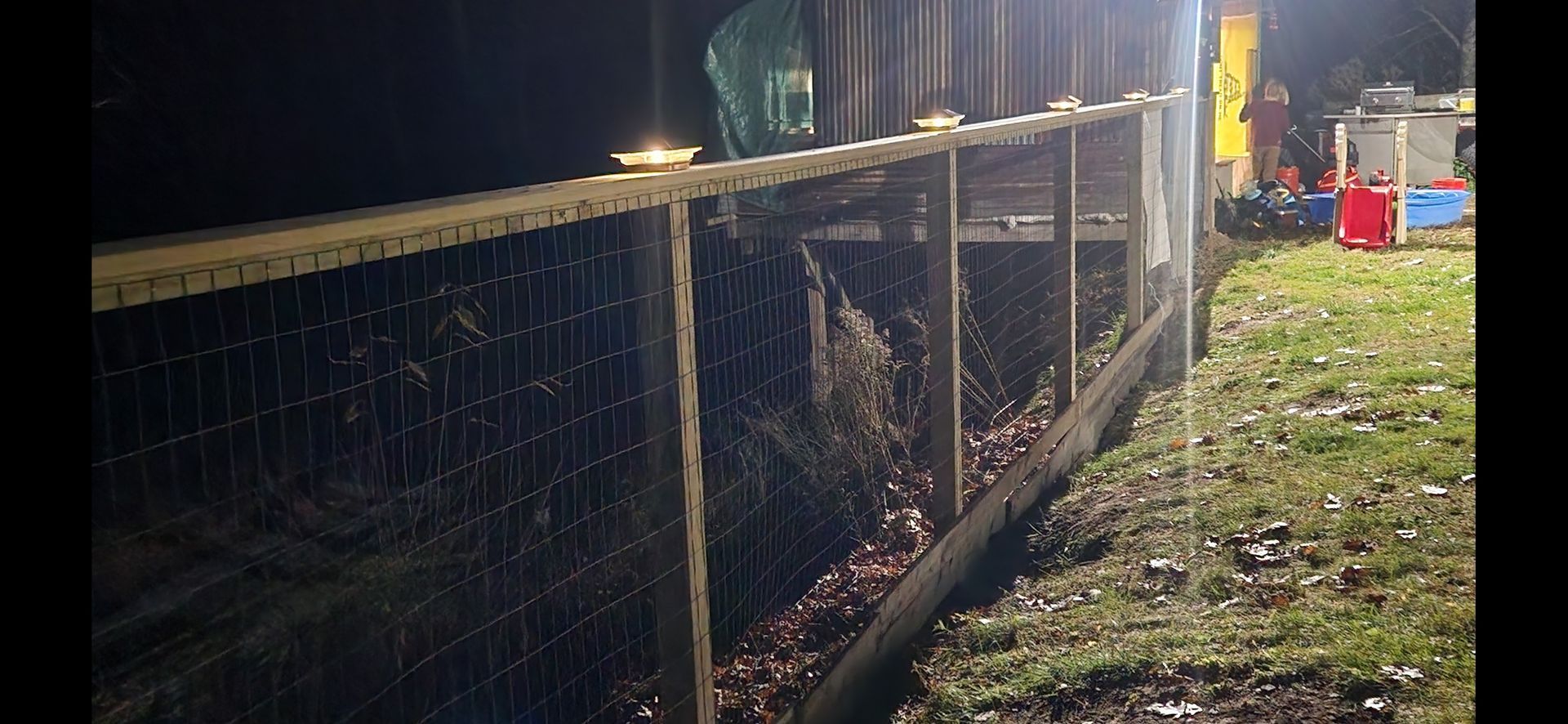Nighttime fenced-off roadside scene with debris, floodlights, and a grassy verge