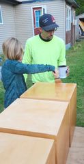 Child handing a mug to a man beside stacked wooden boxes outdoors near a house