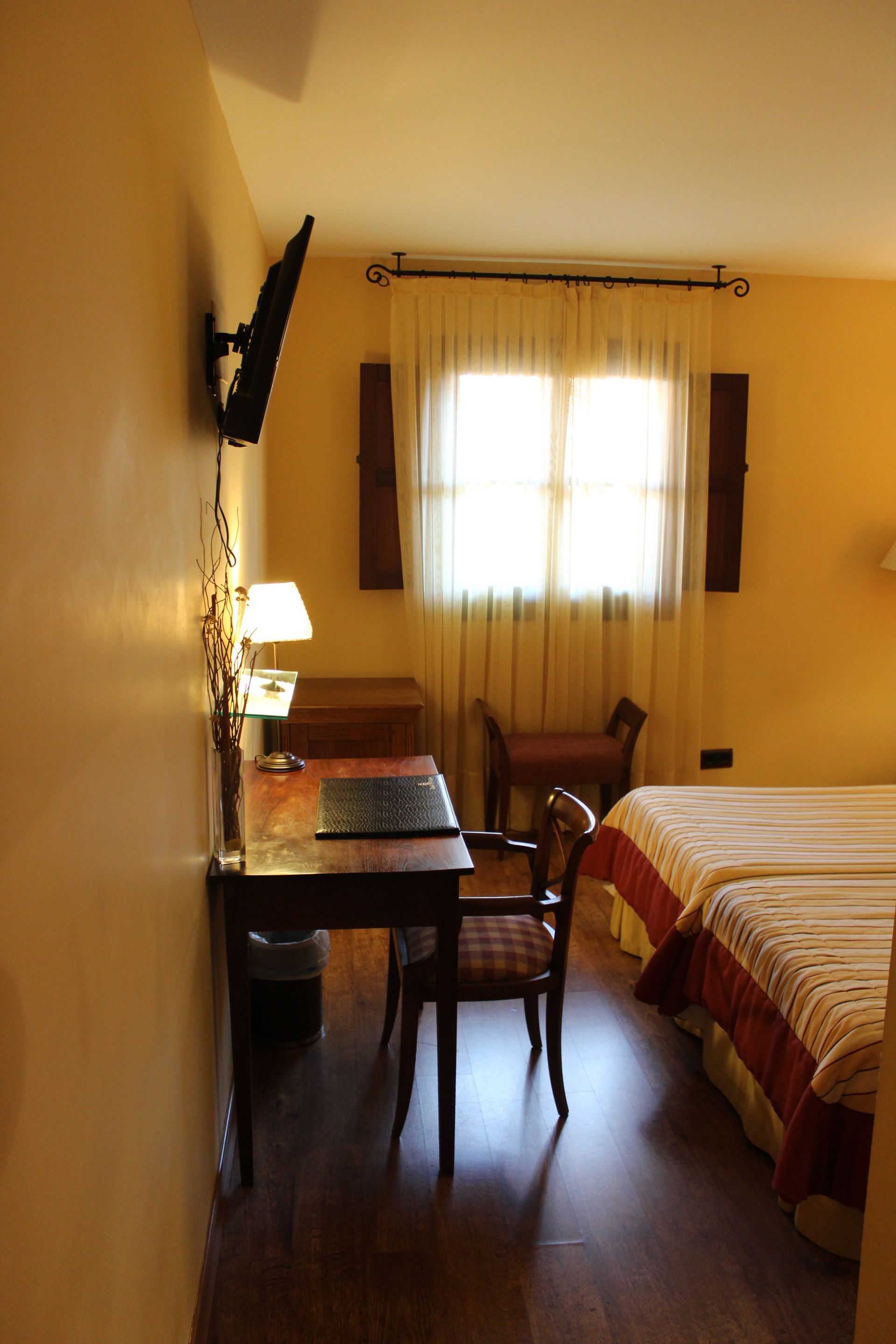 Habitación pequeña con cama, escritorio, silla y ventana con cortinas de luz amarilla cálida.
