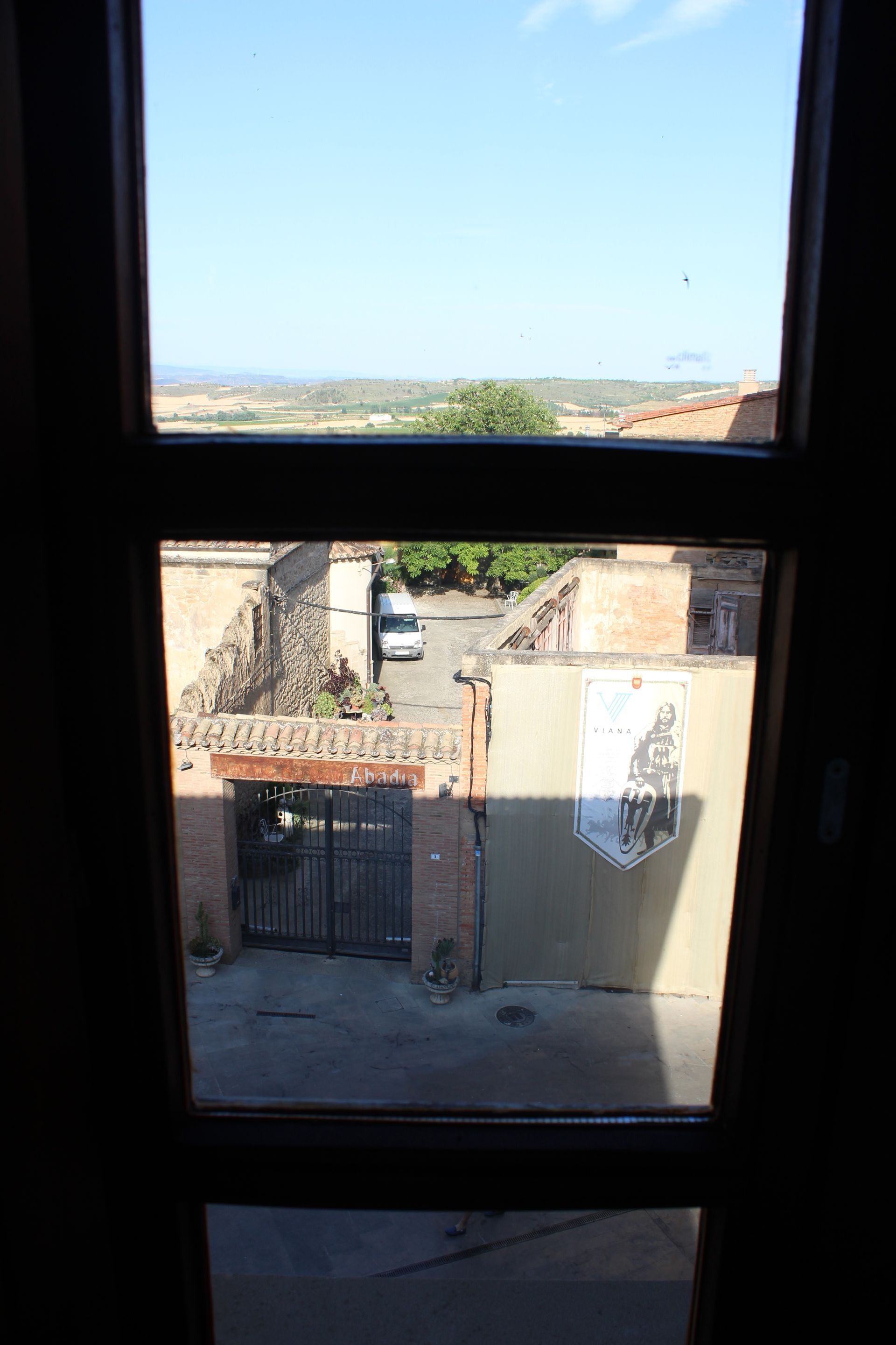 Vista a través de una ventana hacia un patio soleado y edificios de piedra bajo un cielo azul despejado.