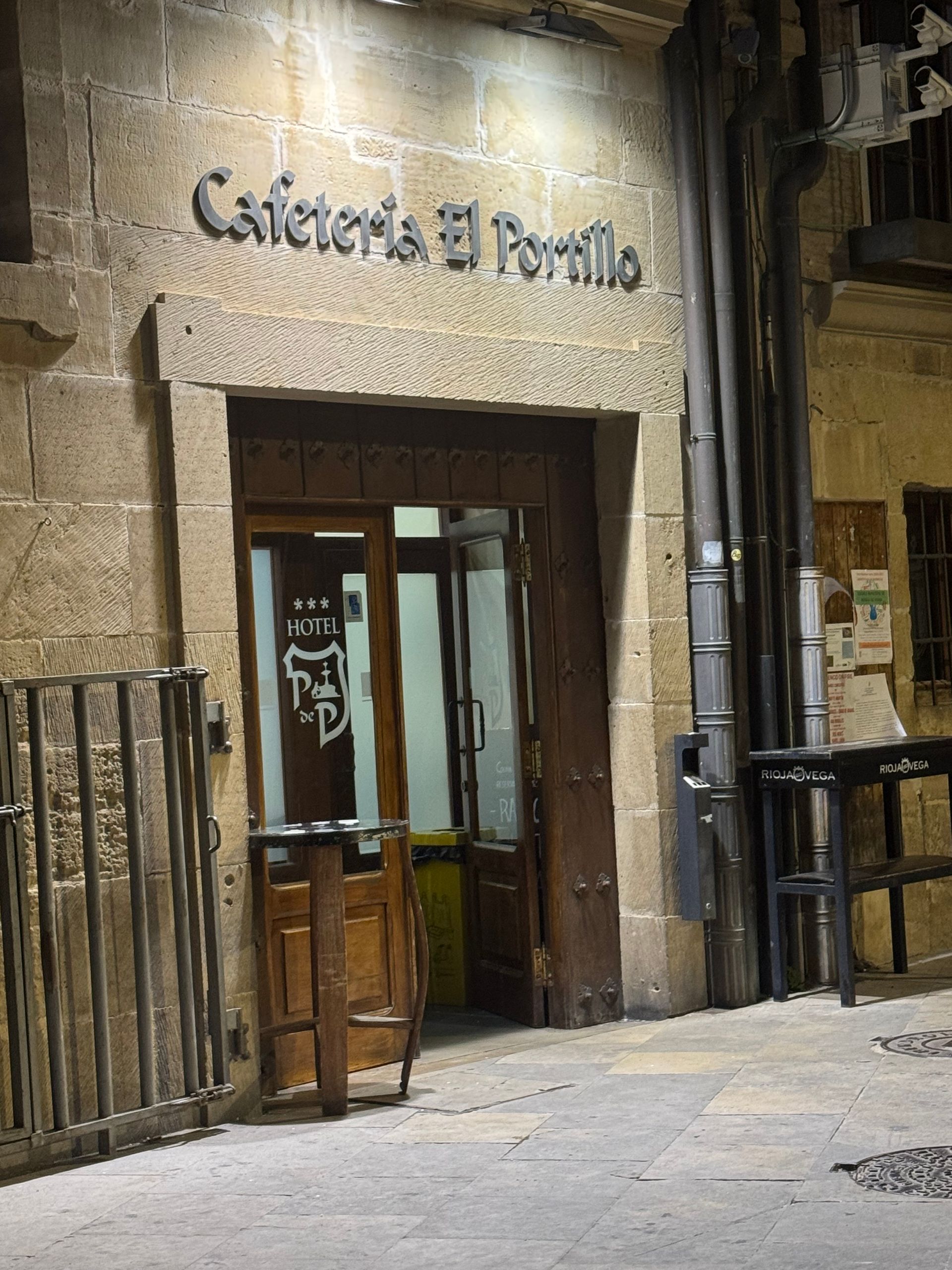 Entrada a la cafetería El Portillo en una calle empedrada, con una puerta de madera abierta y barandillas de hierro.