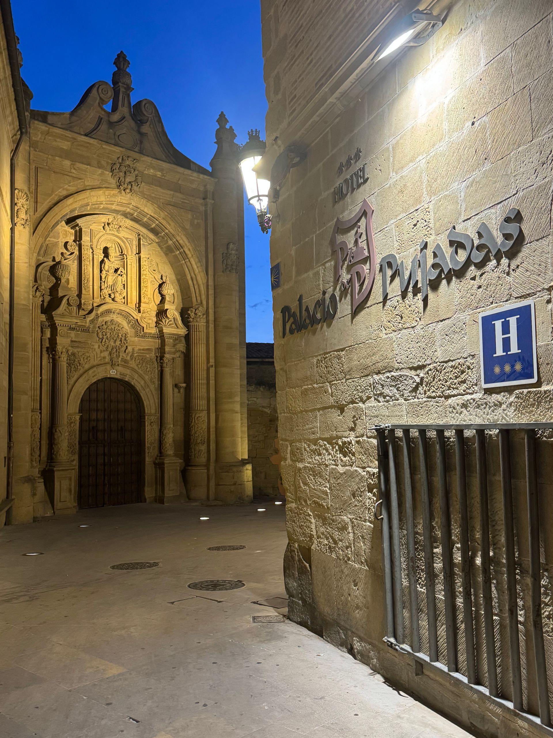 Patio histórico de piedra al anochecer con ventanas iluminadas, farolas cálidas y algunas personas paseando.