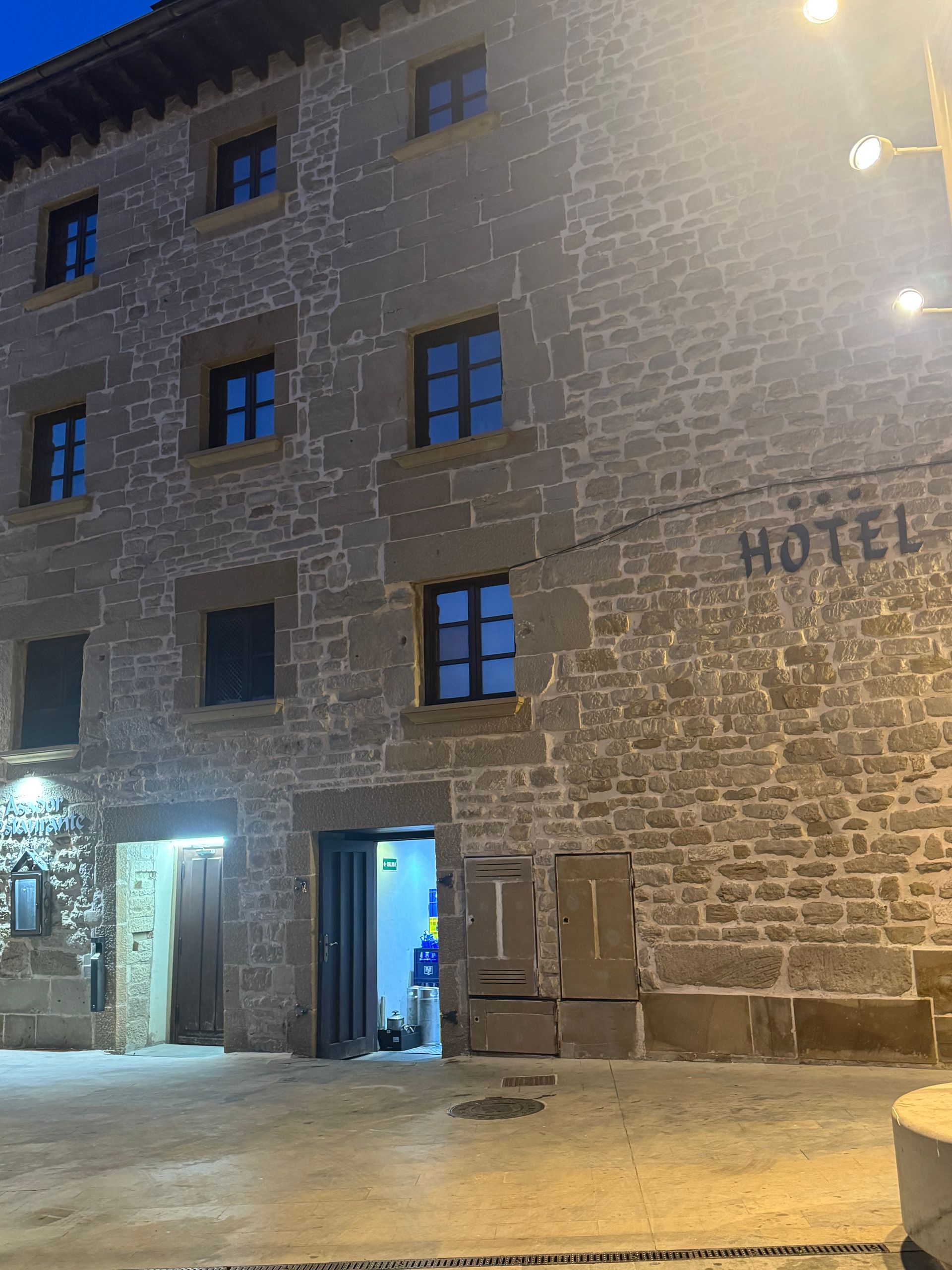 Fachada de un hotel de piedra nevada por la noche, con ventanas iluminadas y puerta de entrada iluminada con luz azul.