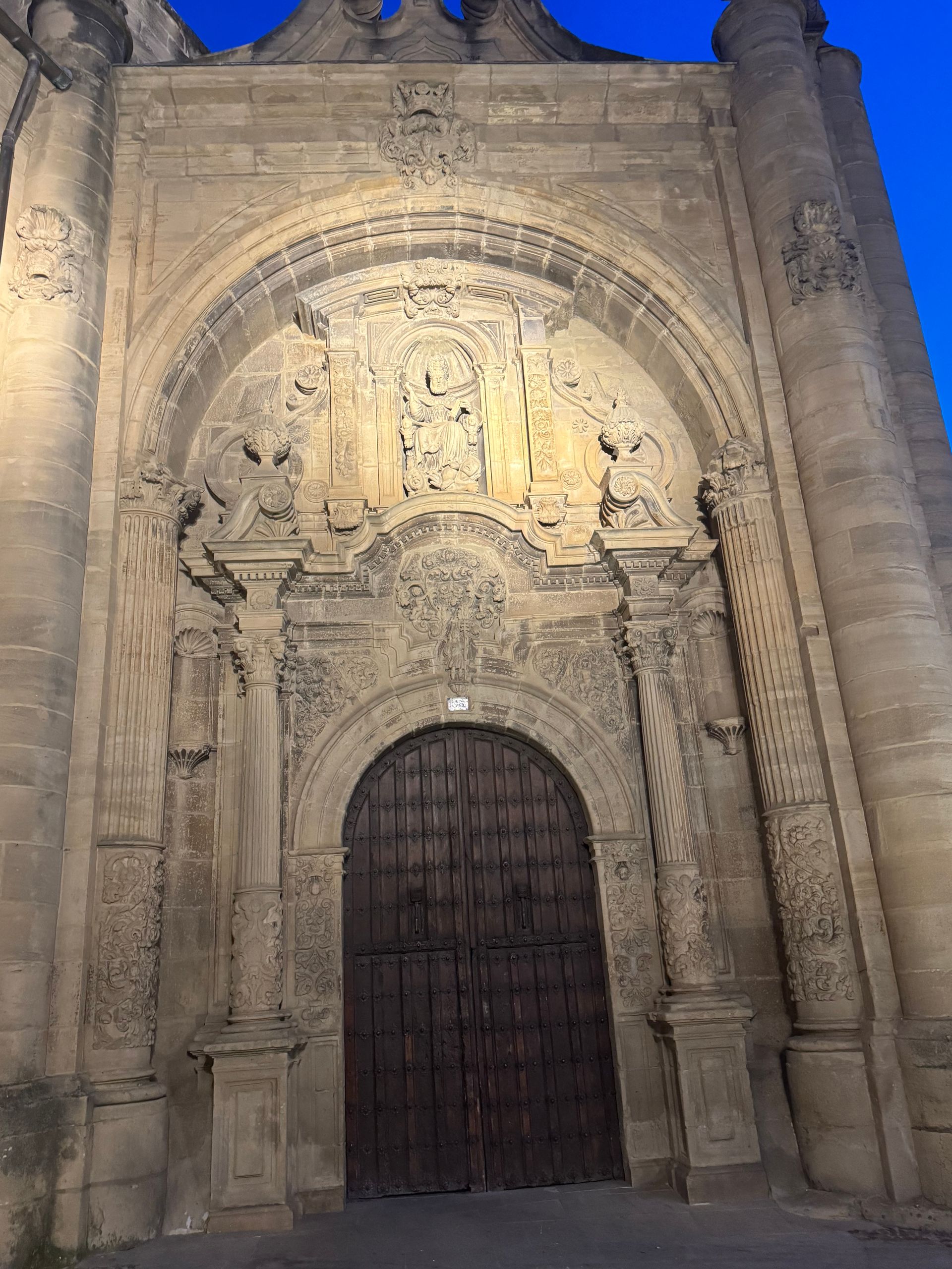 Portal de iglesia de piedra ornamentado con arco tallado y puerta de madera cerrada, iluminado contra un cielo azul.