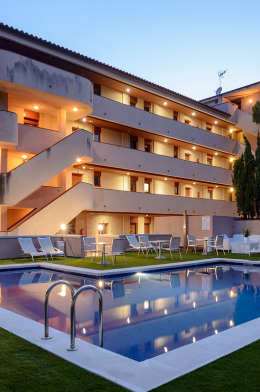 Edificio de apartamentos con balcones iluminados junto a una piscina exterior reflectante al atardecer.