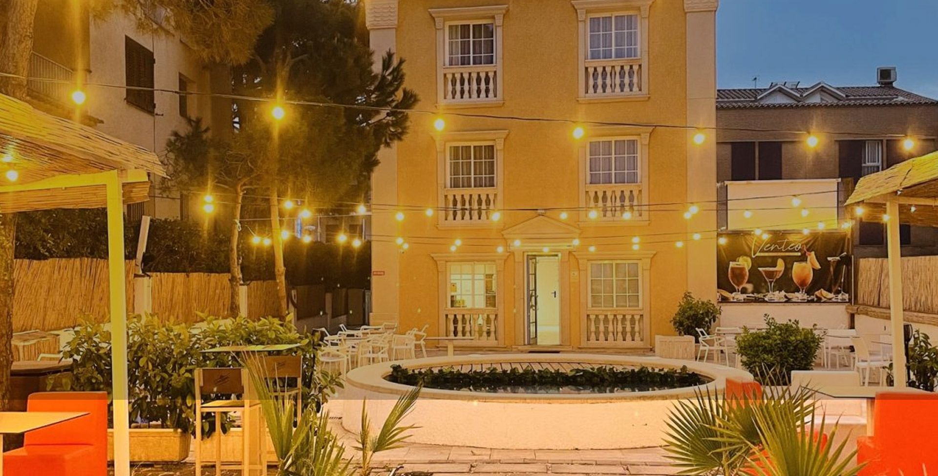 Edificio amarillo iluminado por la noche con un patio, una fuente y asientos de color naranja cálido.