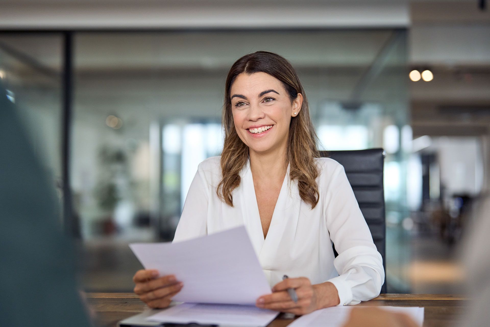 Woman in white blouse smiles, holding papers, at a table in an office setting.
