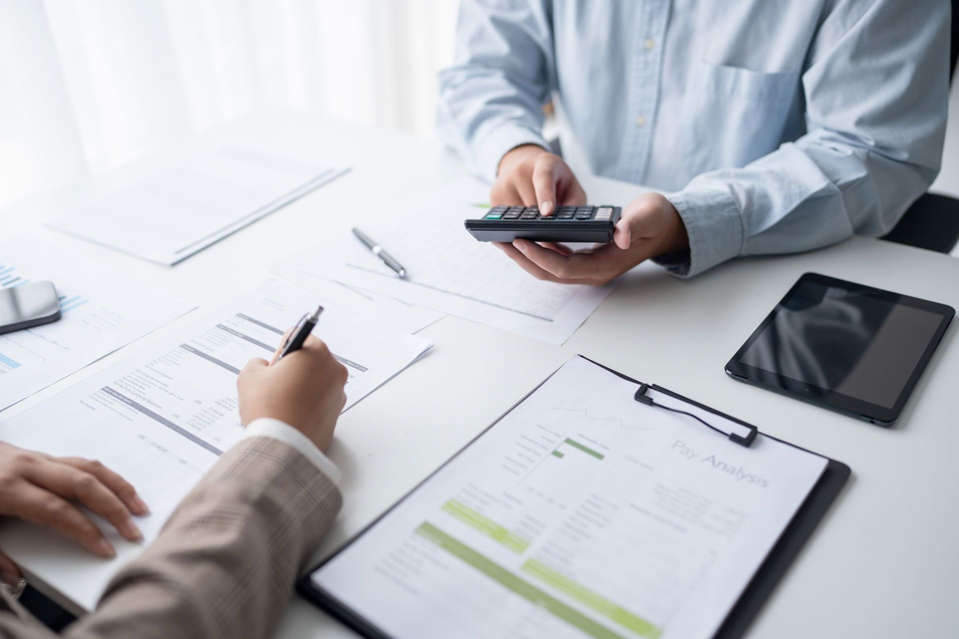 Two people at a desk review documents and use a calculator, likely discussing finances.
