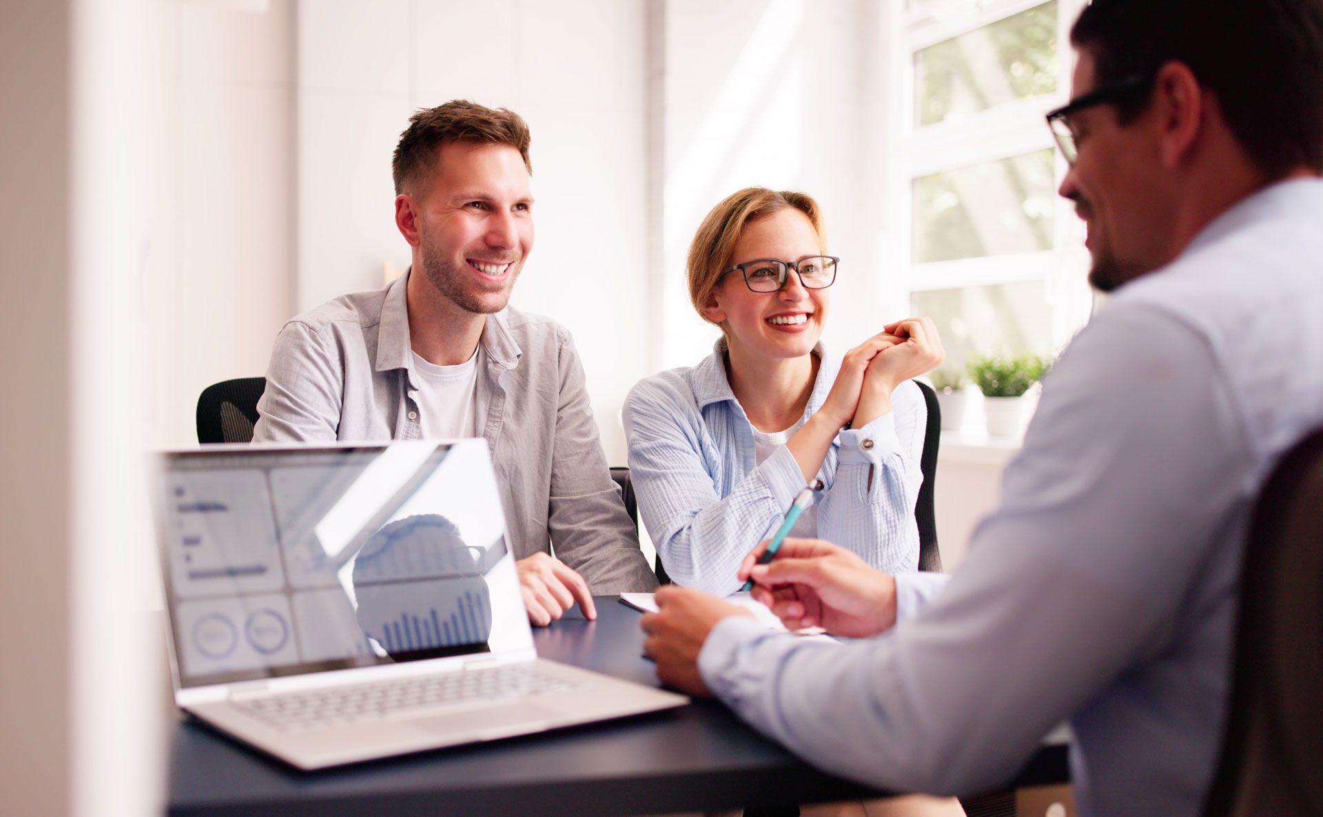 Couple smiles at a person, sitting at a table with a laptop. Meeting inside a bright office.