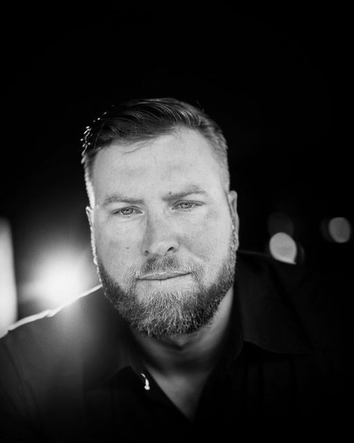 Man with a beard, looking at the viewer, against a dark background, lit from below.