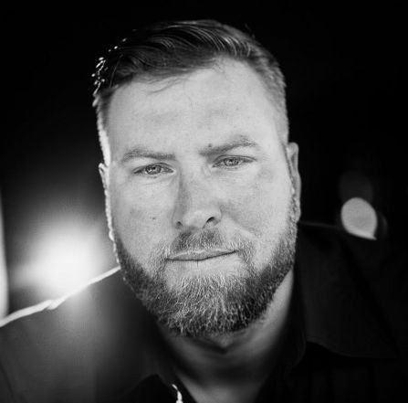 Man with a beard looks directly at the camera in a close-up shot; black and white photo.