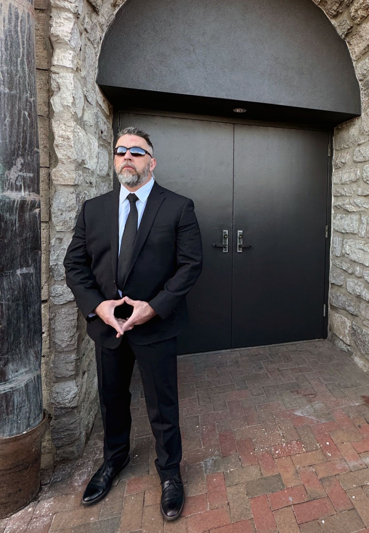 Man in black suit and sunglasses stands in front of black door, arms crossed, on brick path.