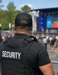 Security guard in black uniform at outdoor concert, seen from behind near barricades.
