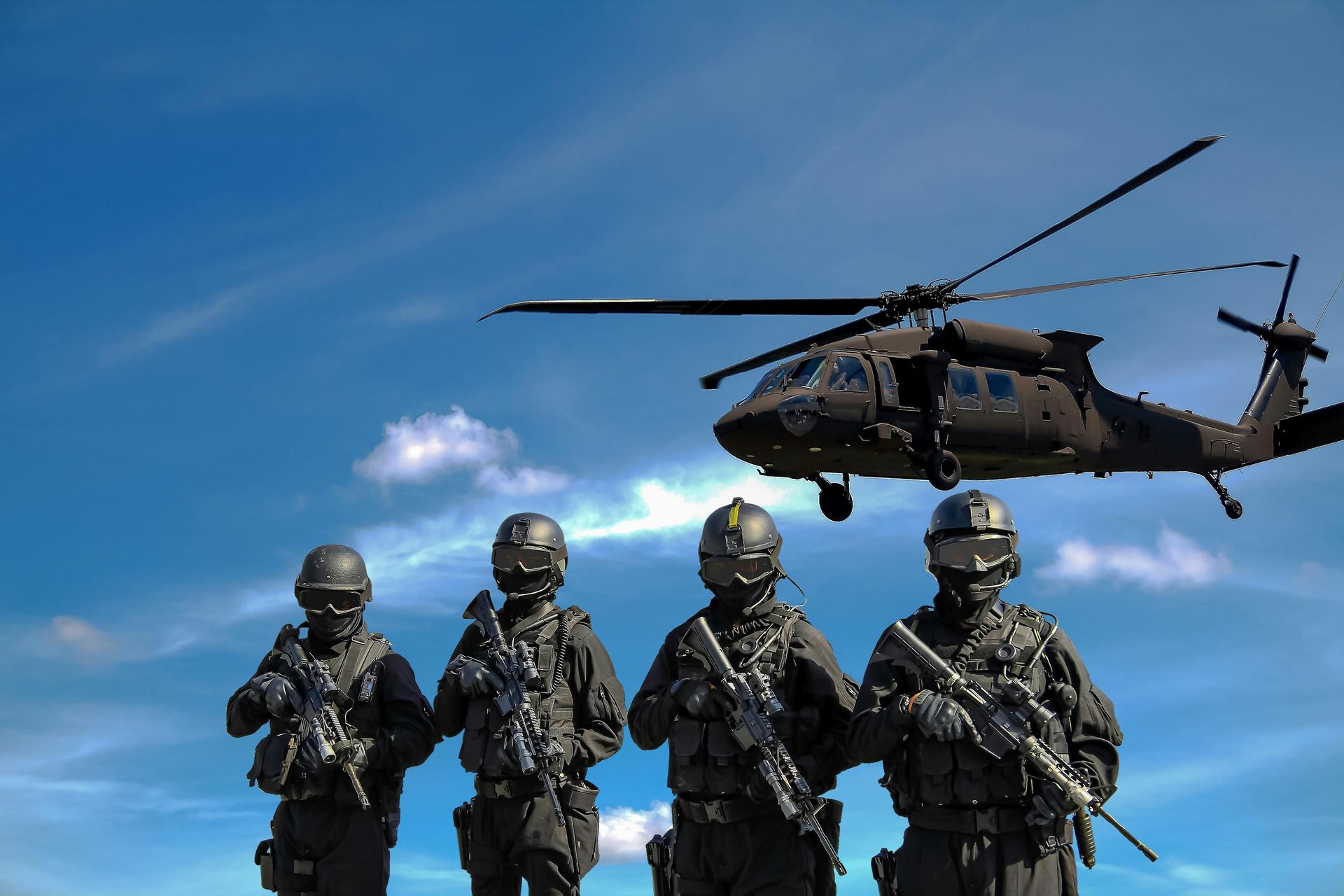 Four soldiers in tactical gear with rifles, standing in front of a helicopter against a blue sky.