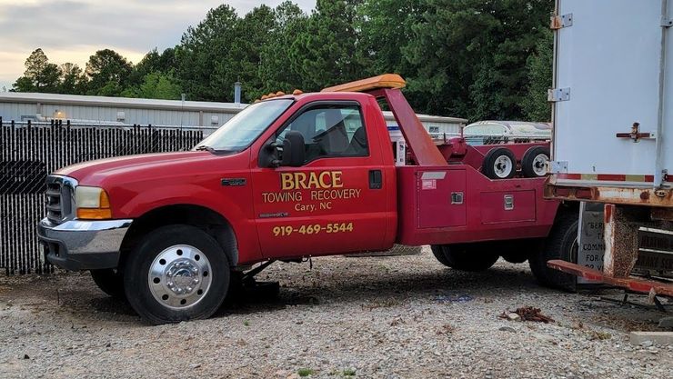 Red tow truck with flatbed hauling a white box truck in a gravel lot
