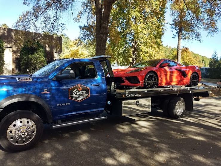 Blue tow truck carrying a red sports car on a flatbed trailer outdoors under trees.