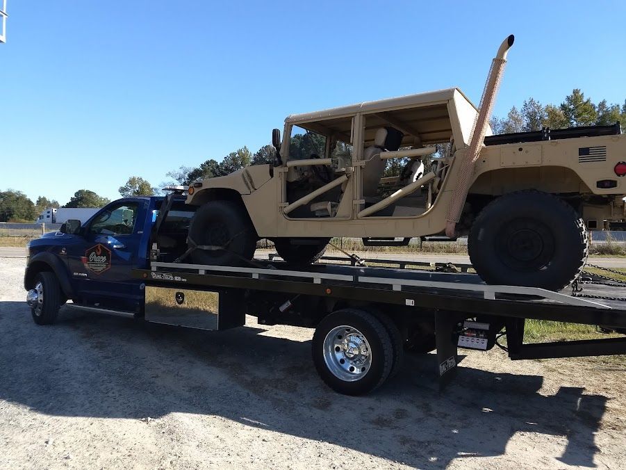 Tow truck hauling a tan military Humvee on a flatbed trailer in a sunny outdoor lot