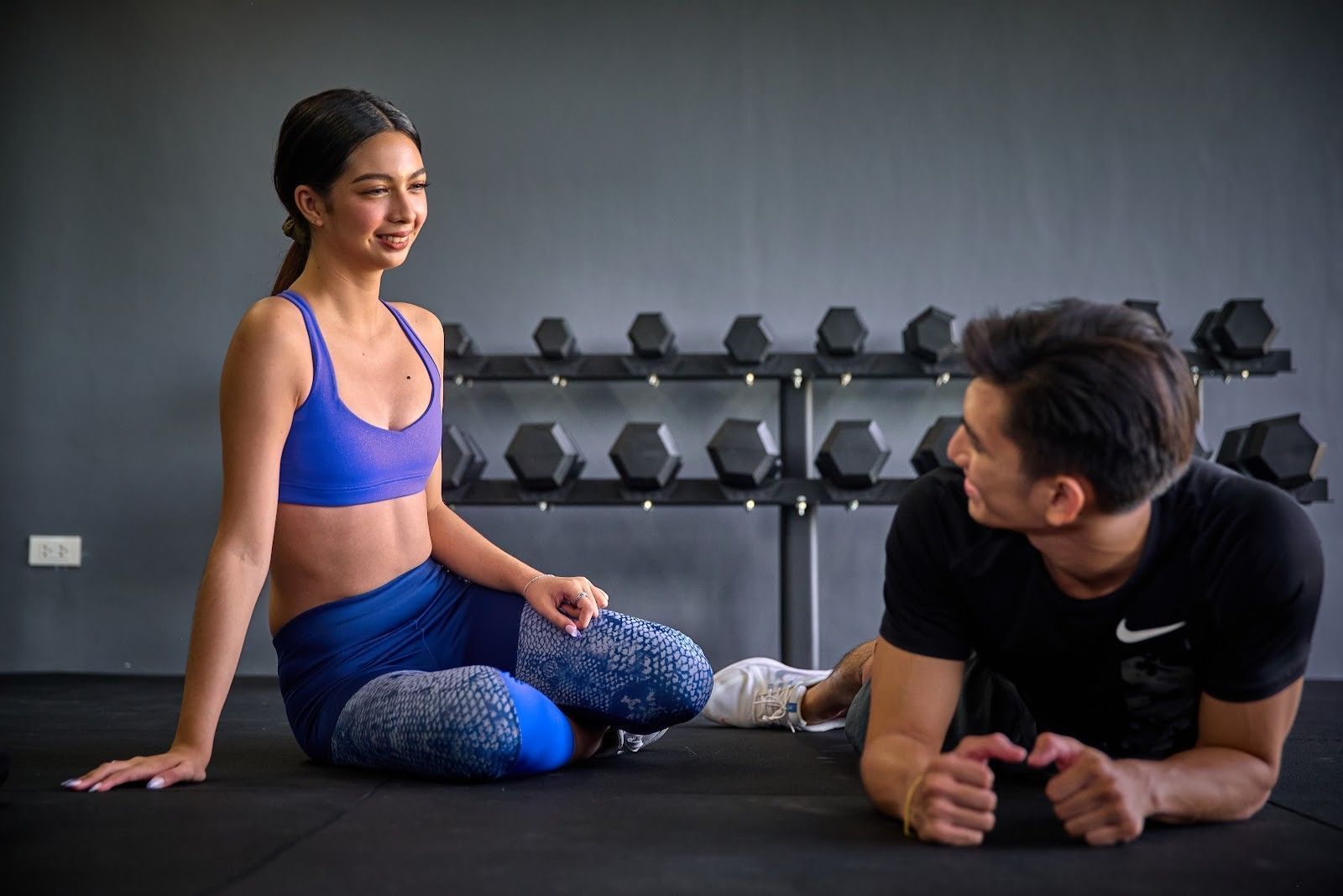 Two people resting on gym mats beside dumbbells, one sitting and smiling while the other lies on his side.