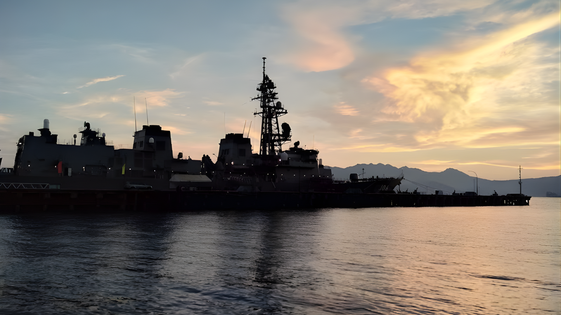 Silhouette of a naval ship at sunset on calm water, with mountains in the distance