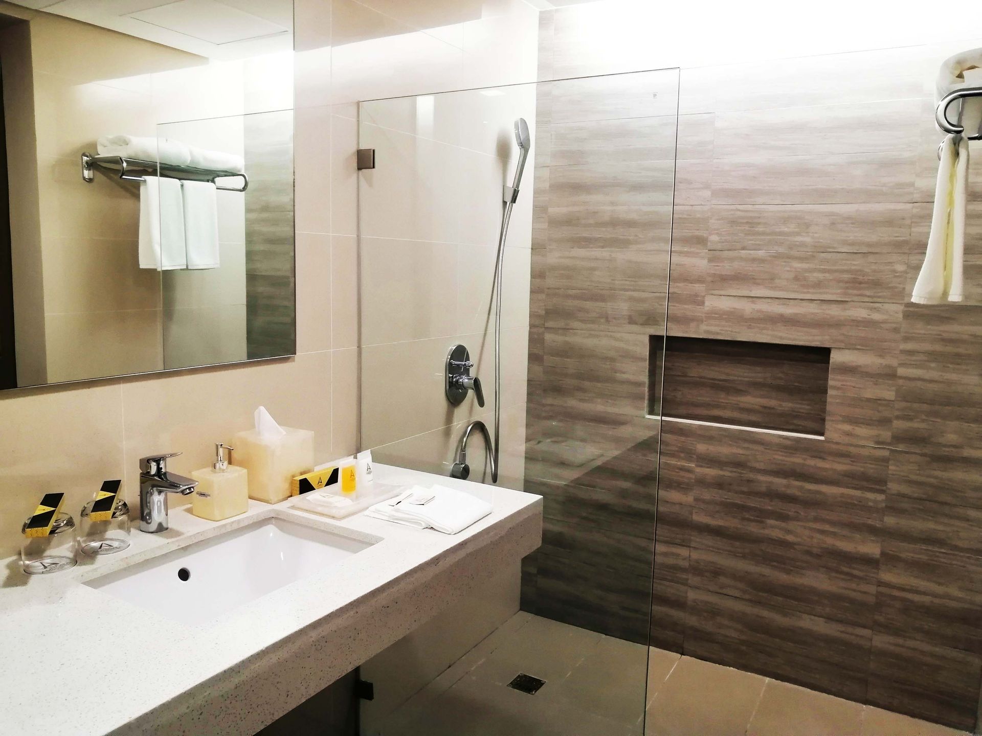 Bathroom with a white sink, mirror, glass shower door, and brown tiled walls. Towels hang on a rack.