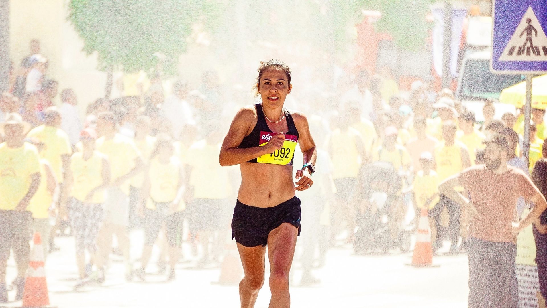 Runner in black shorts and yellow bib runs ahead in a bright, crowded race finish area.