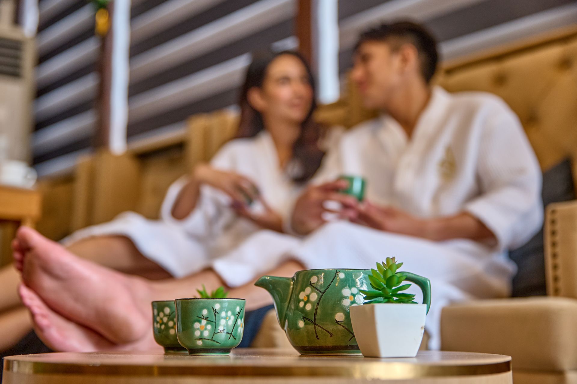 Couple in white robes relaxing and talking in a spa setting with a teapot and cups on a table.