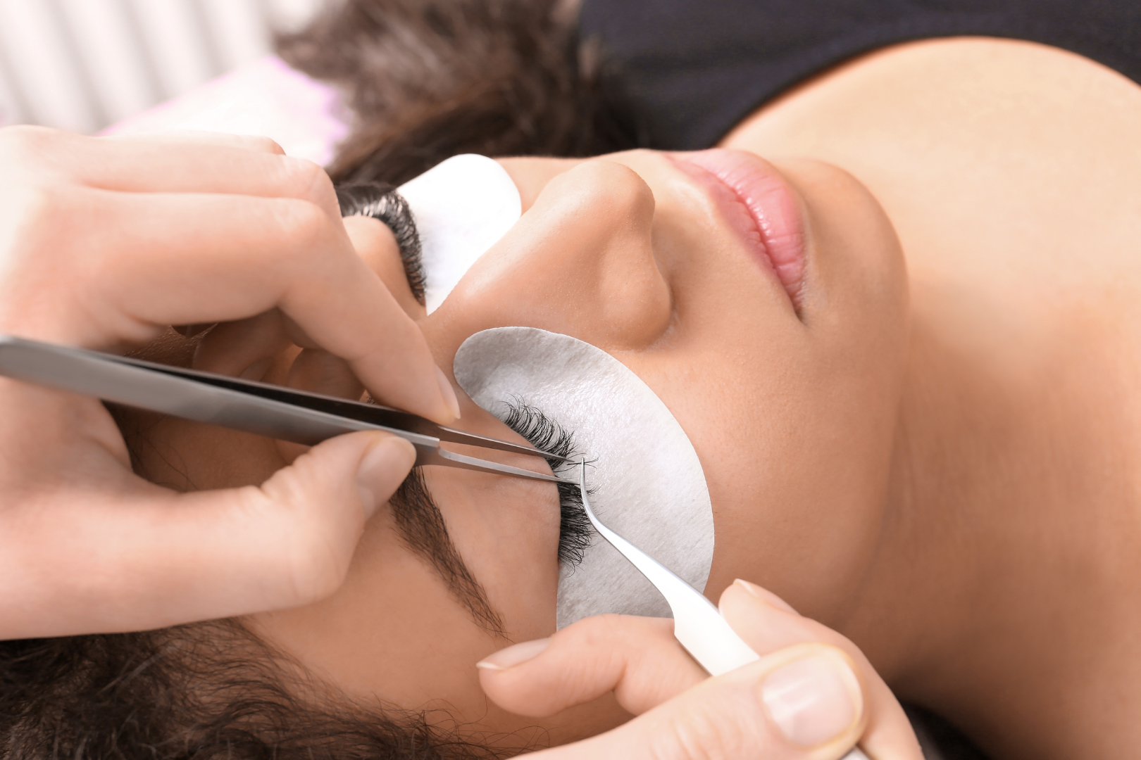 Woman receiving eyelash extensions; technician using tweezers, eye covered with pad.