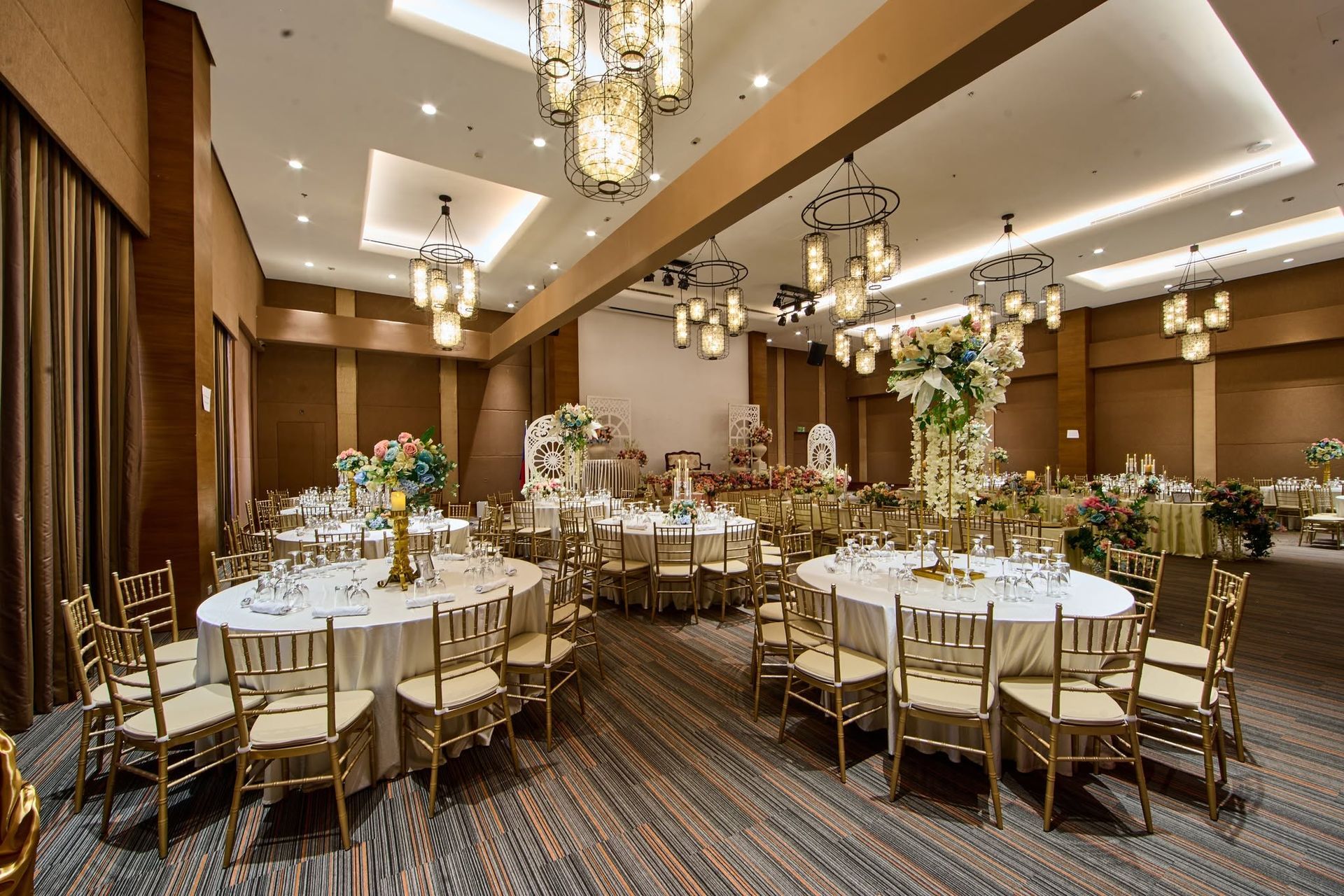Elegant banquet hall with round white-clothed tables, gold chairs, chandeliers, and patterned carpet