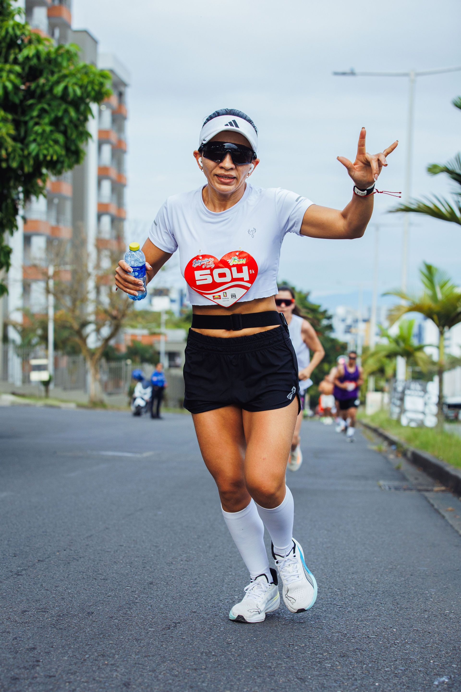 Runner in white shirt and black shorts smiling on a city street during a race, holding a bottle and flashing a peace sign