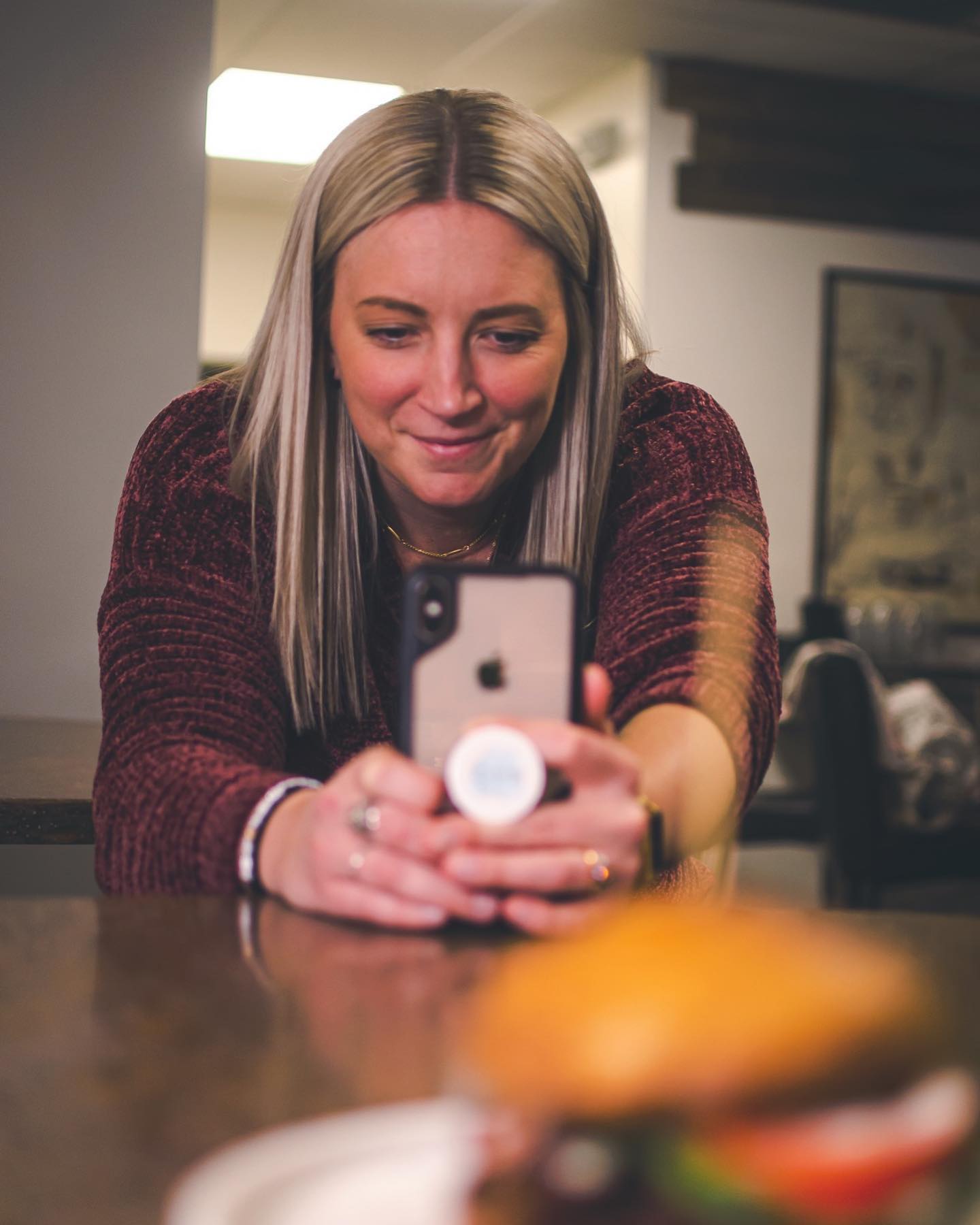 Woman takes a photo of a burger with her phone, smiling. She has blonde hair, wearing a burgundy sweater, indoors.