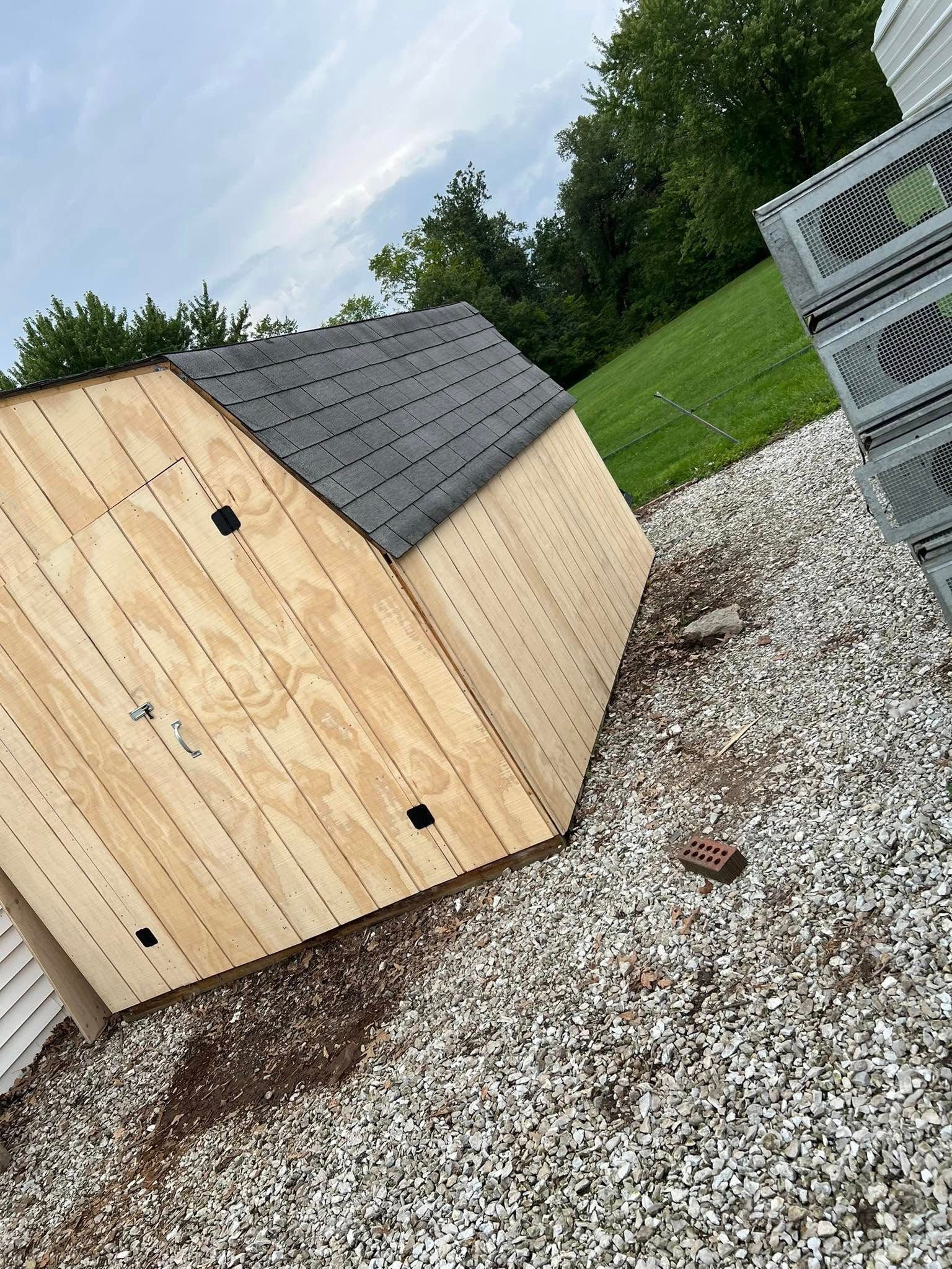 Small wooden shed with a dark shingled roof on a gravel yard beside green grass and stacked wire crates.