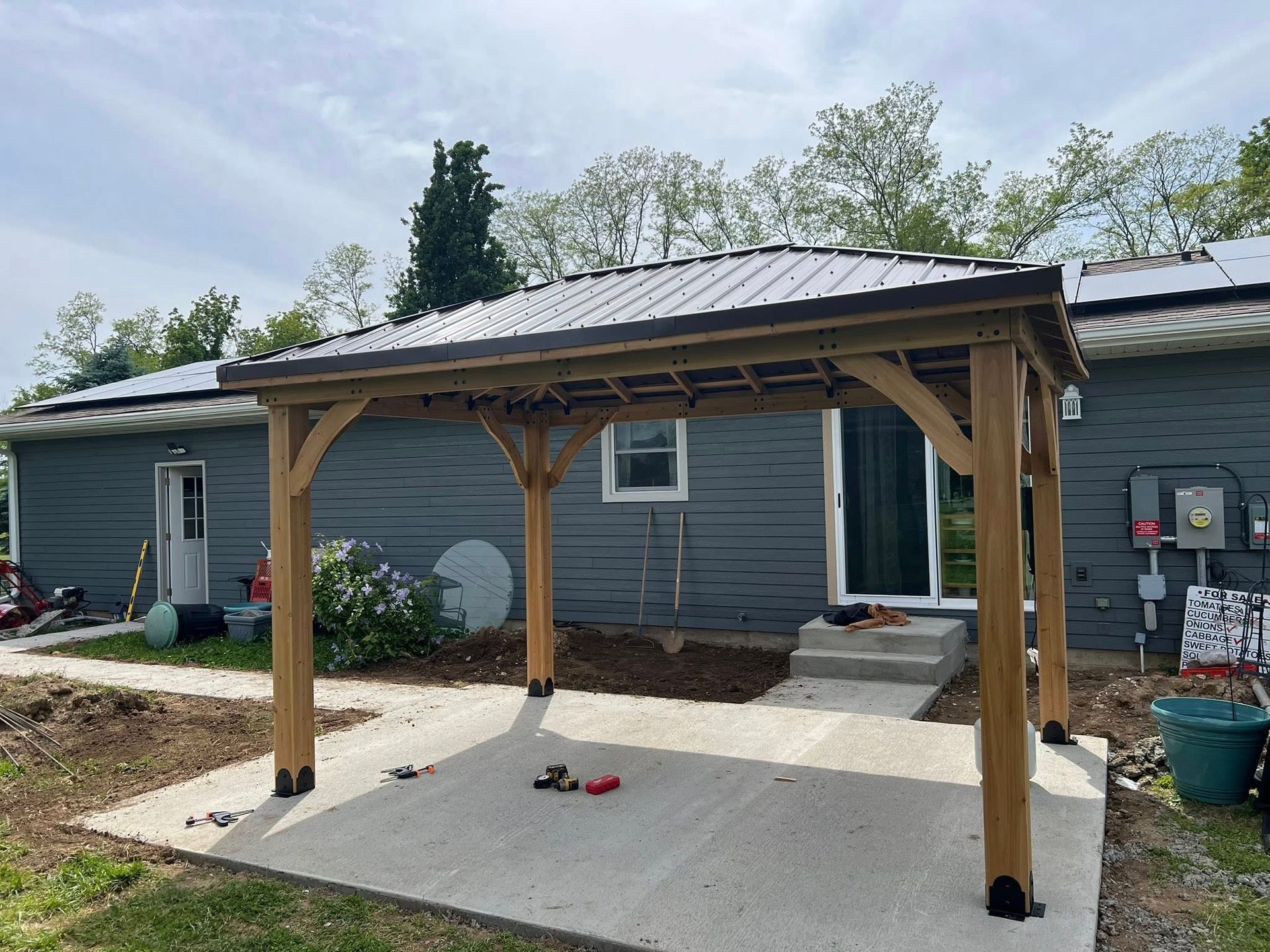 Wooden pergola with a metal roof on a concrete patio behind a gray house.