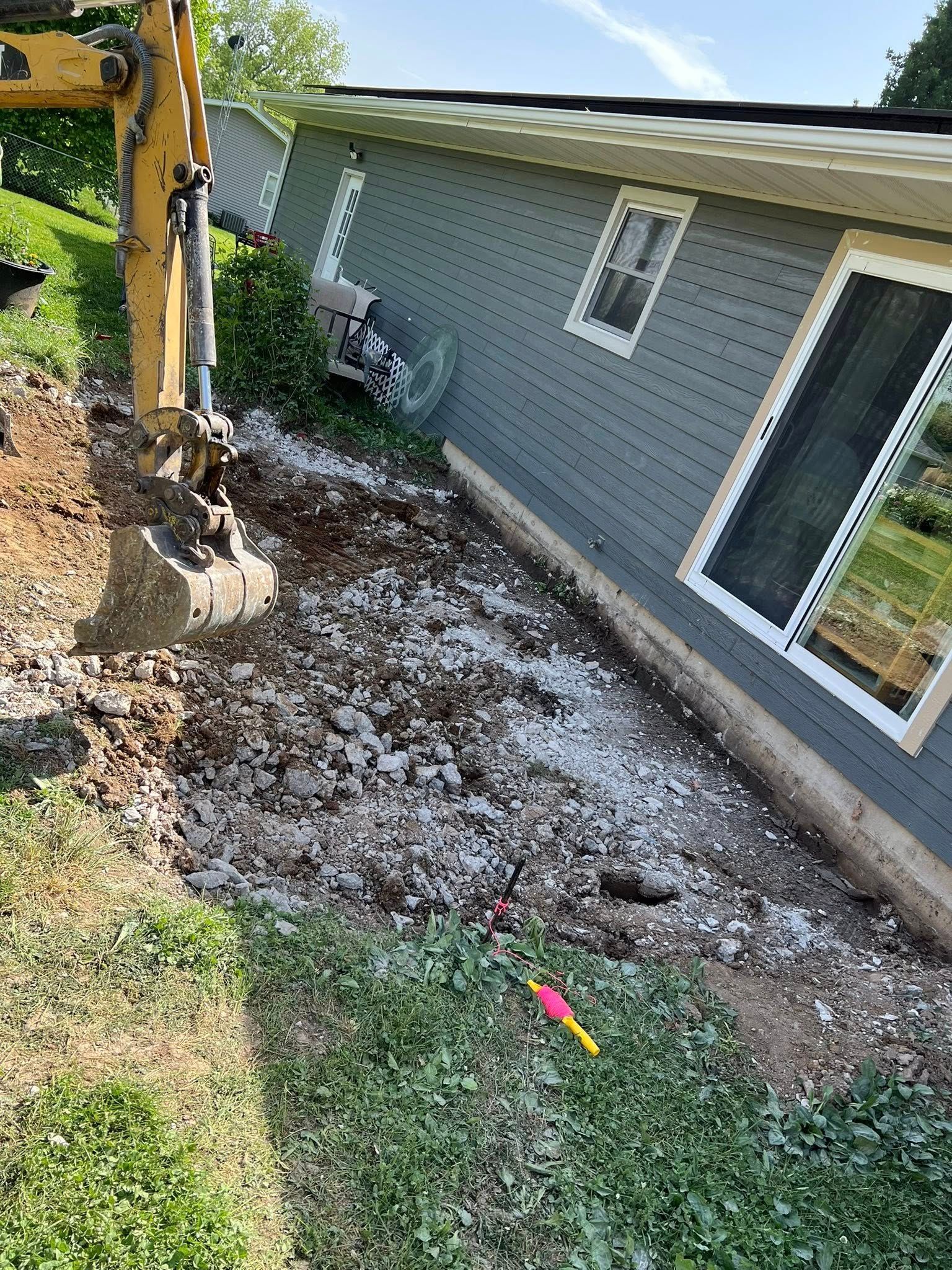 Excavator beside a house, clearing dirt and debris along the foundation near windows and a sliding glass door.