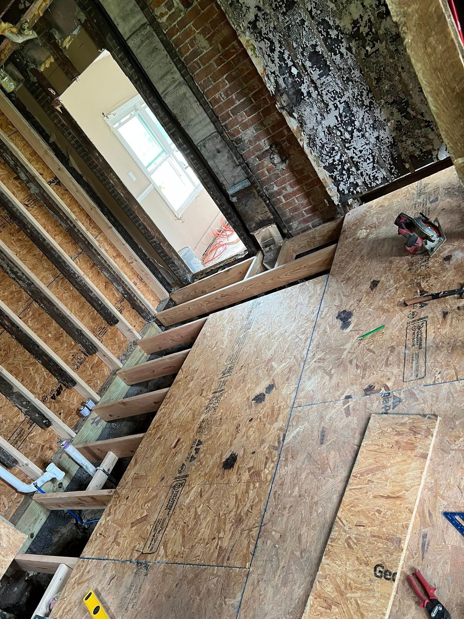 Interior of a stripped room with exposed wall studs, plywood floor, and a window at the far end.